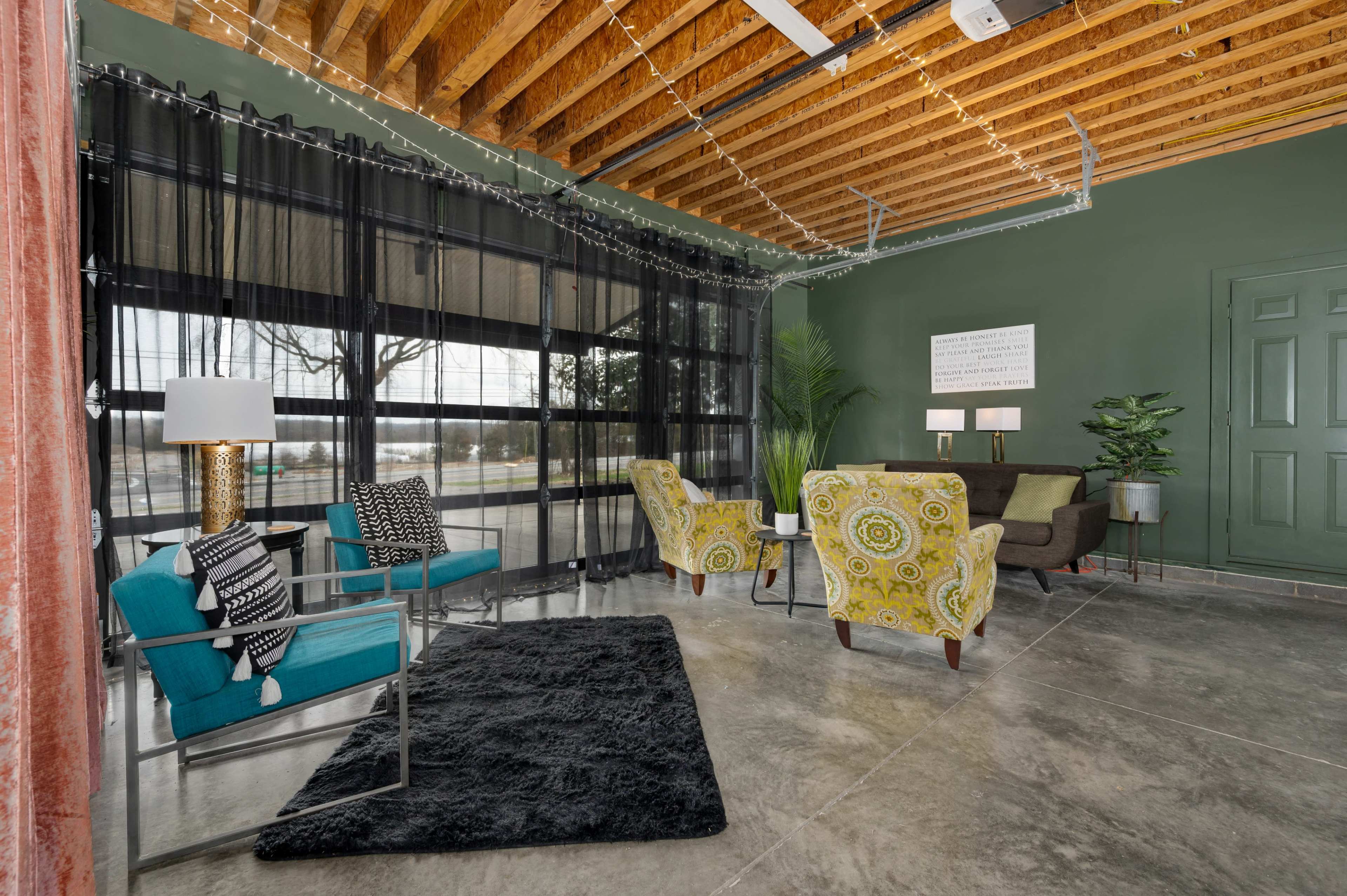 A cozy interior space featuring a mix of modern seating arrangements, a black rug, and plants, with large windows adorned by sheer curtains.