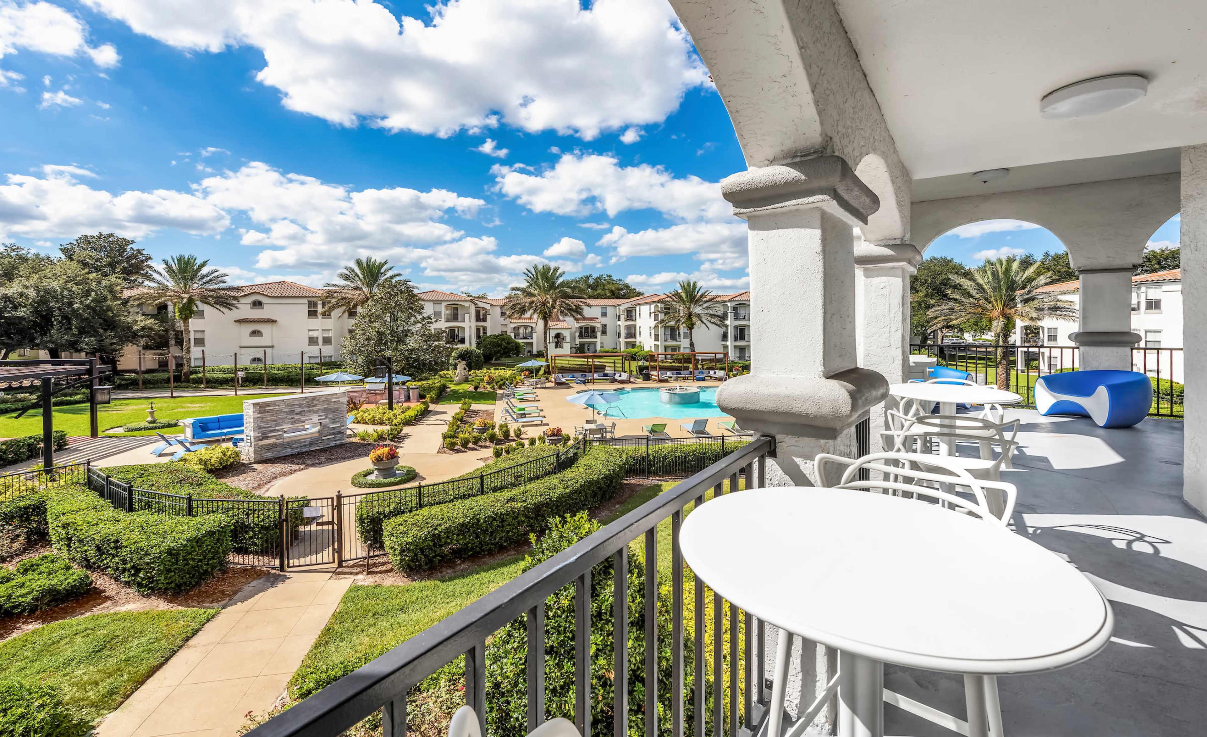 The image shows a balcony view overlooking a landscaped pool area surrounded by palm trees and residential buildings.