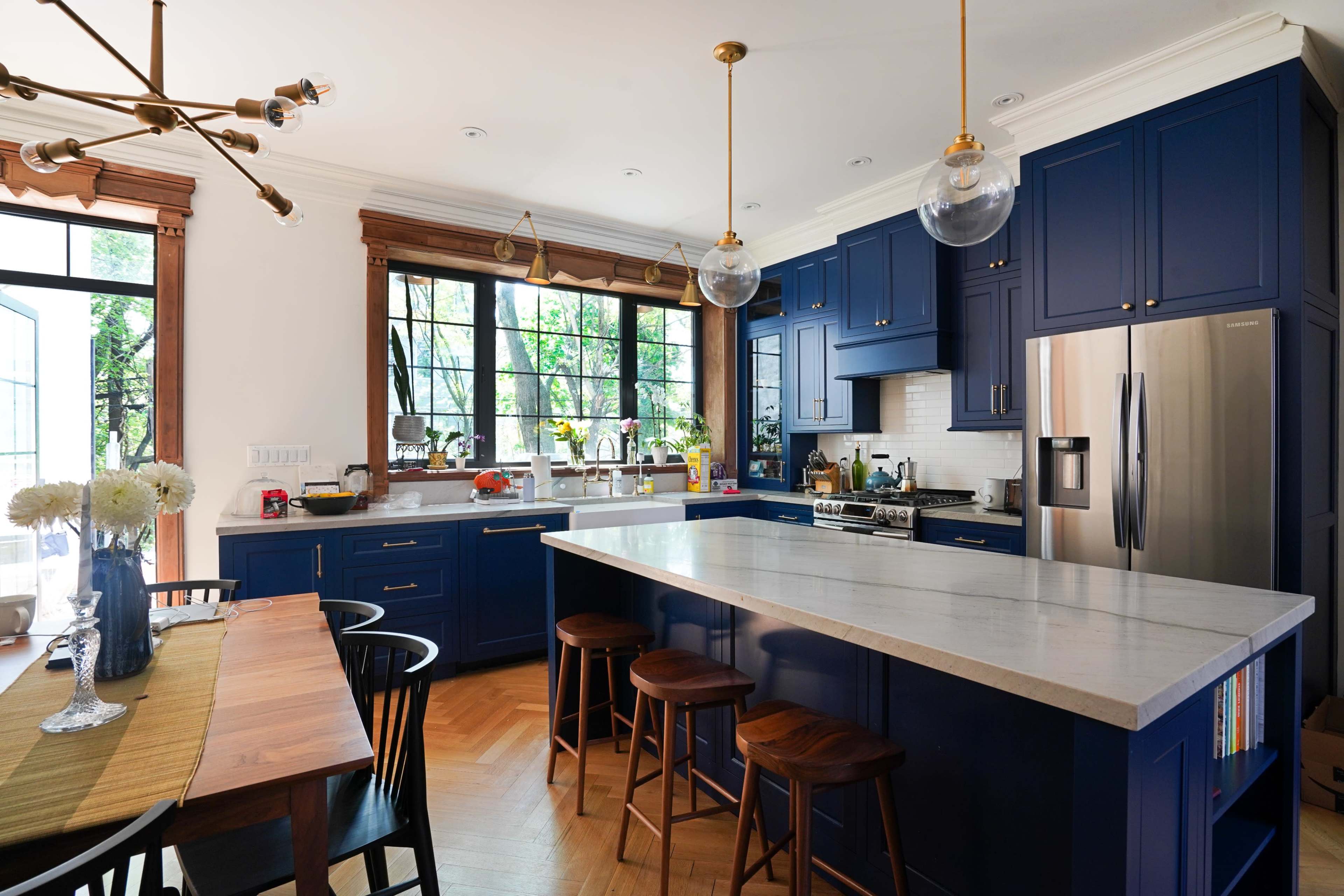 The image shows a modern kitchen with blue cabinetry, a large marble island, and a wooden dining table set against a backdrop of large windows.