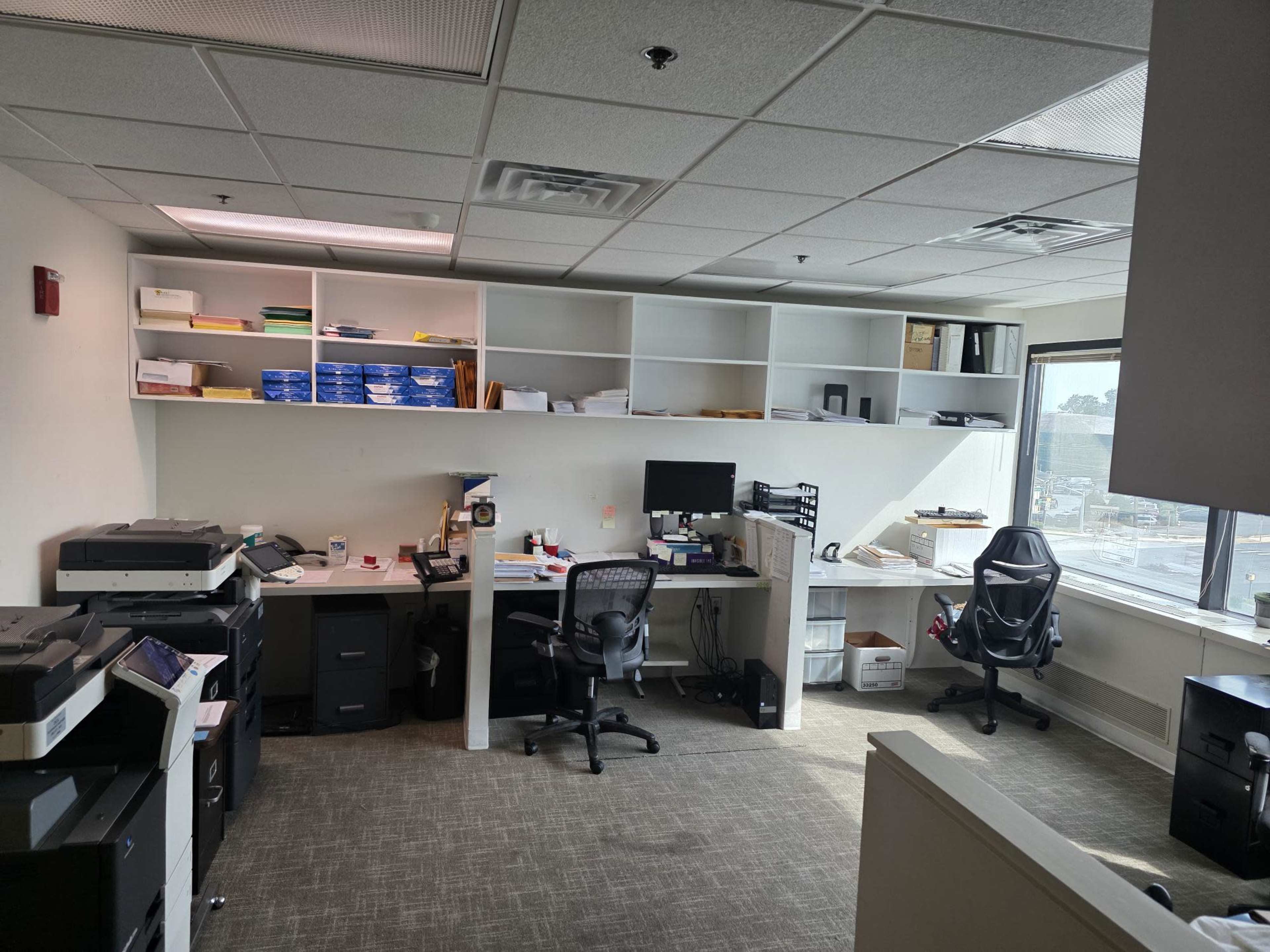 The image shows an office space with desks, printers, and shelving that contains various office supplies.