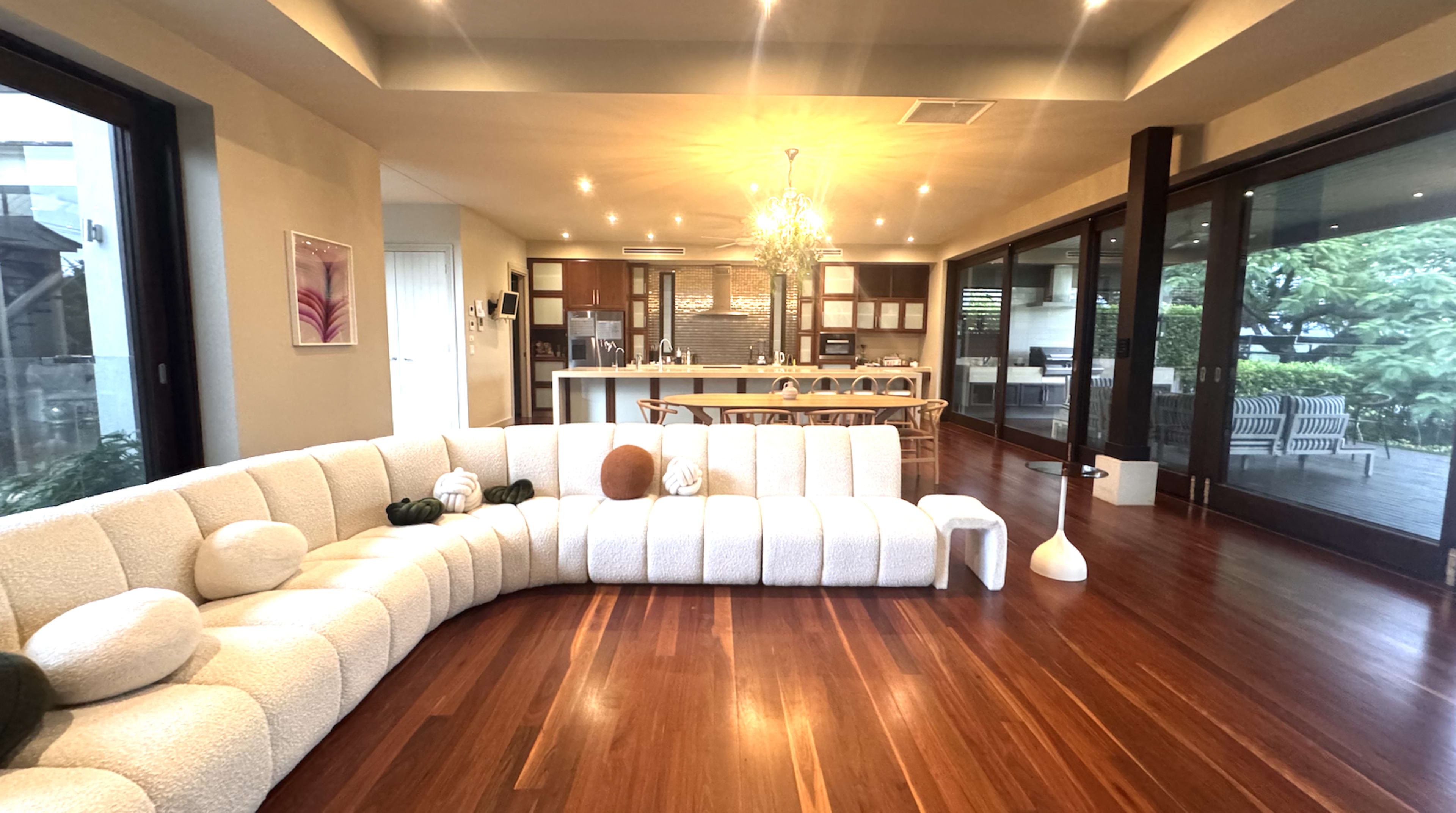 The image shows a modern living space featuring a curved white couch, wooden flooring, and large windows with a view of greenery outside.