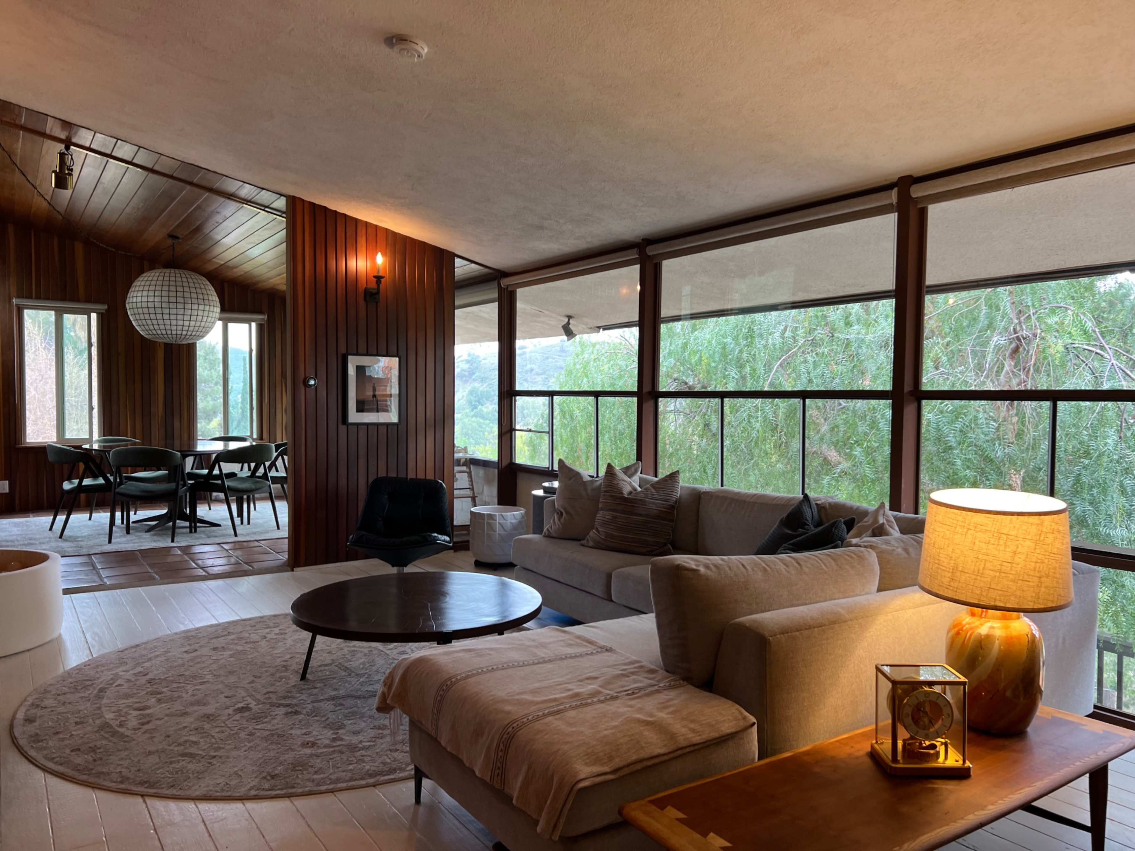 The image shows a modern, open-concept living area with a sectional sofa, round coffee table, and a warm lamp, overlooking a scenic view through large windows.