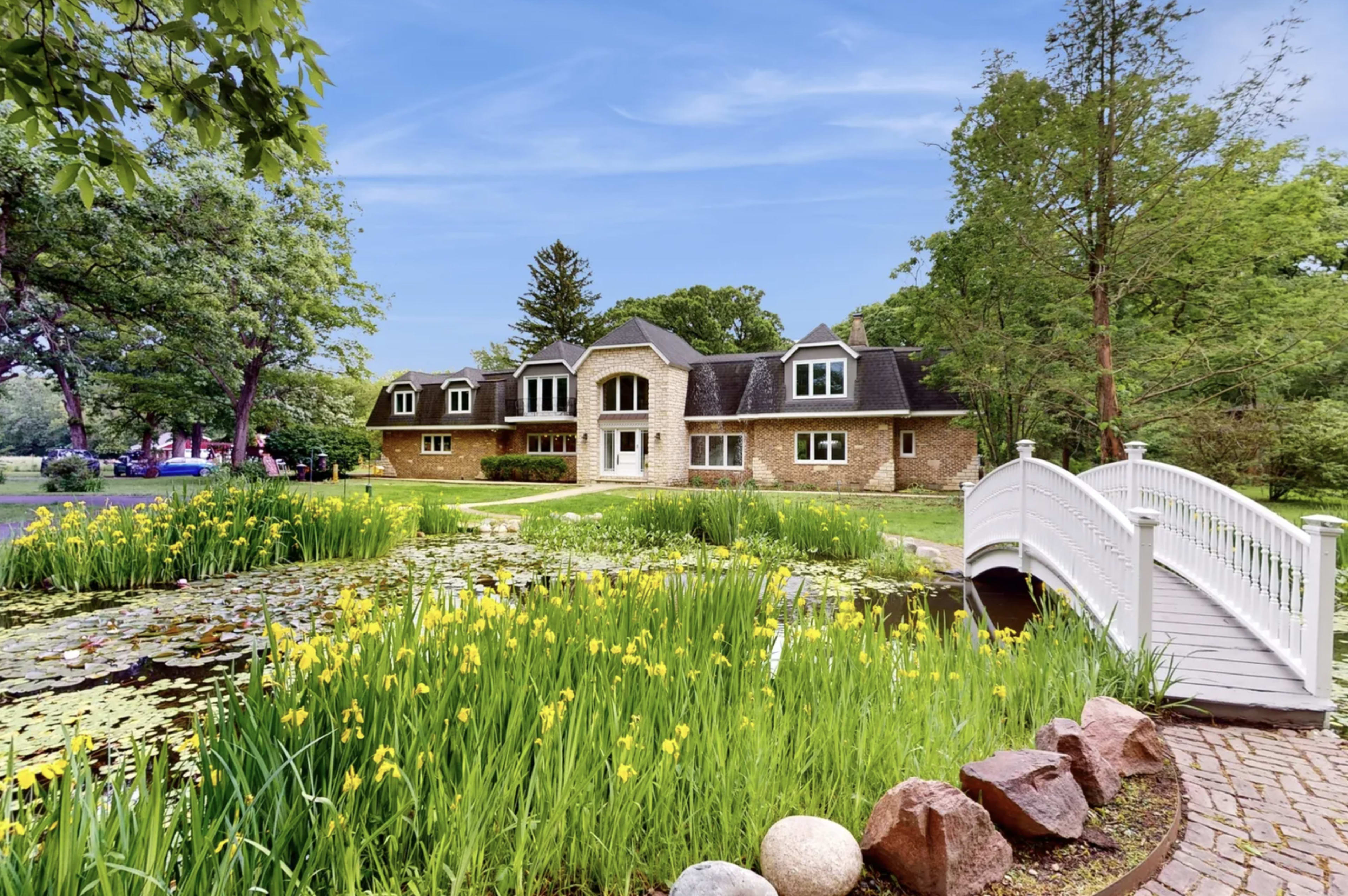 A stone house with multiple gabled roofs is situated near a pond surrounded by greenery and yellow flowers, with a white wooden bridge in the foreground.