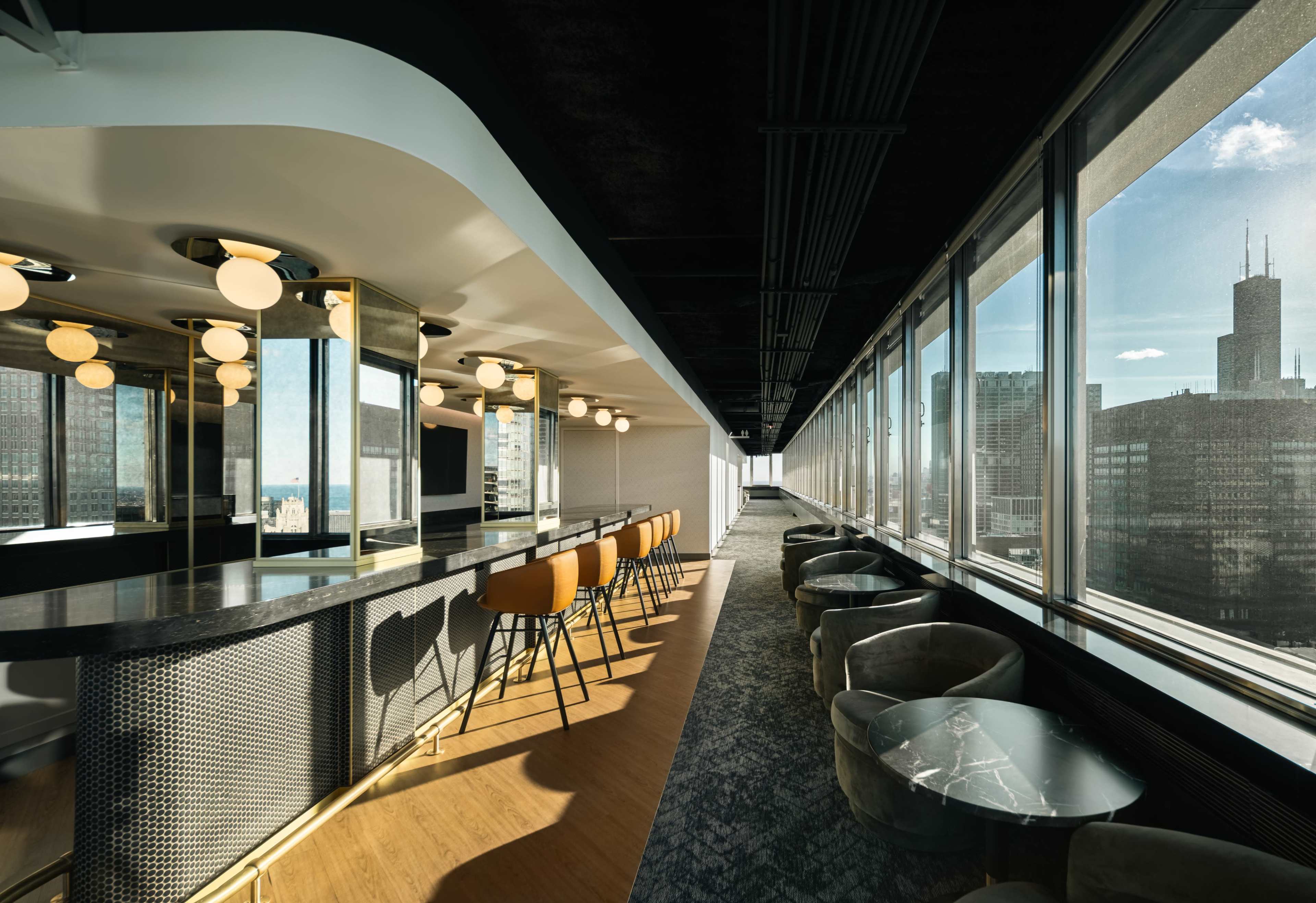 The image shows a modern office space featuring a long bar with orange stools, surrounded by large windows offering a view of a city skyline.
