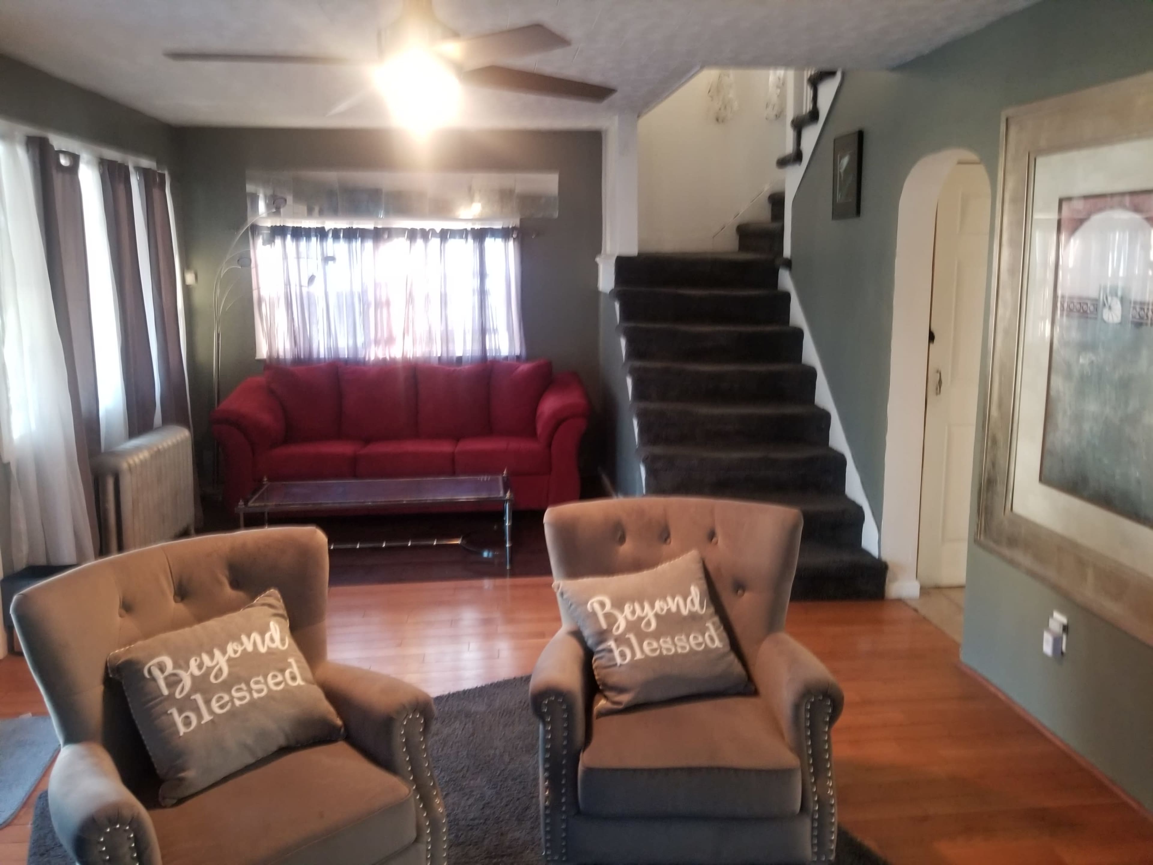 A living room features a red couch, two gray armchairs with decorative pillows, and a staircase leading to an upper level.