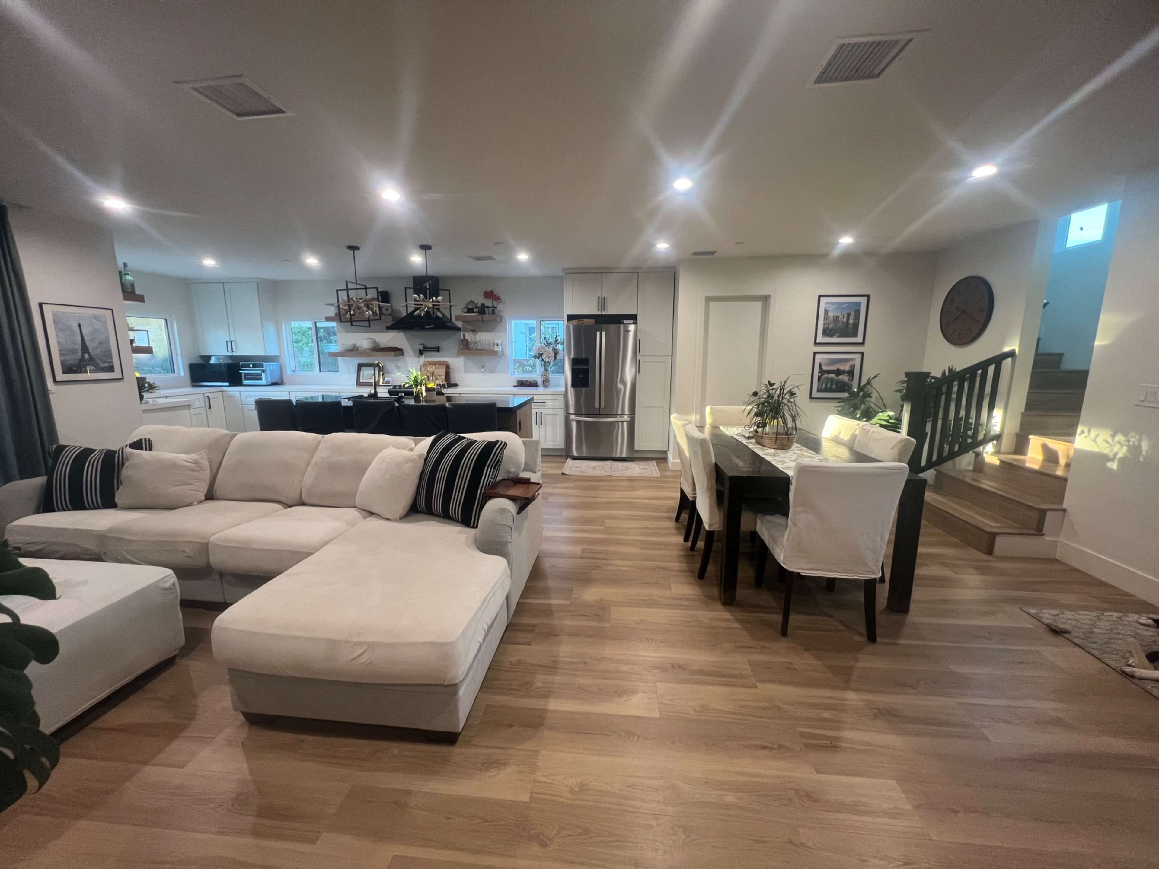 The image shows a modern open-concept living and dining area with a large sectional sofa, a dining table, and a kitchen in the background.