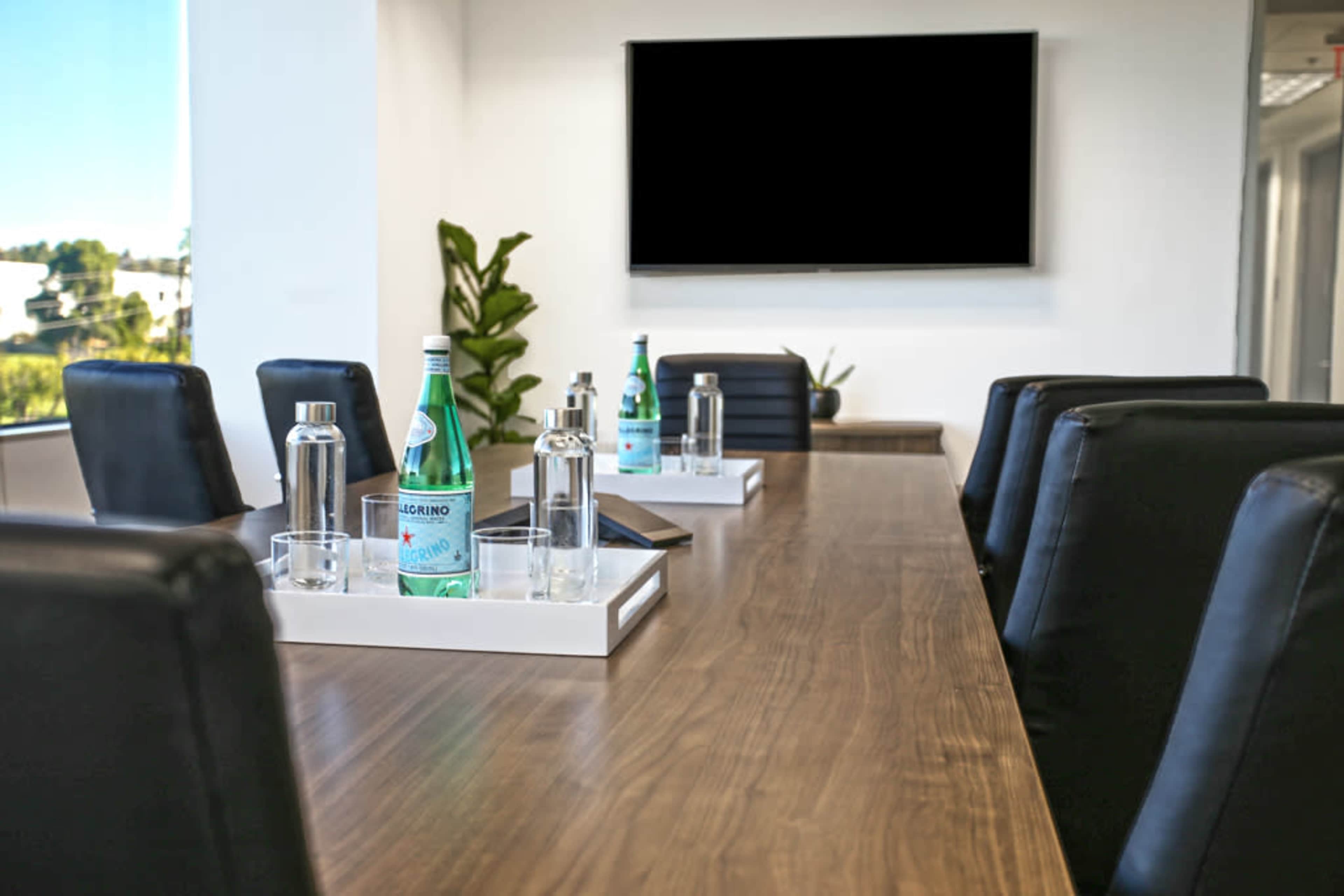 A modern conference room features a long wooden table with bottled water and glasses arranged on trays, flanked by black chairs and a wall-mounted screen.
