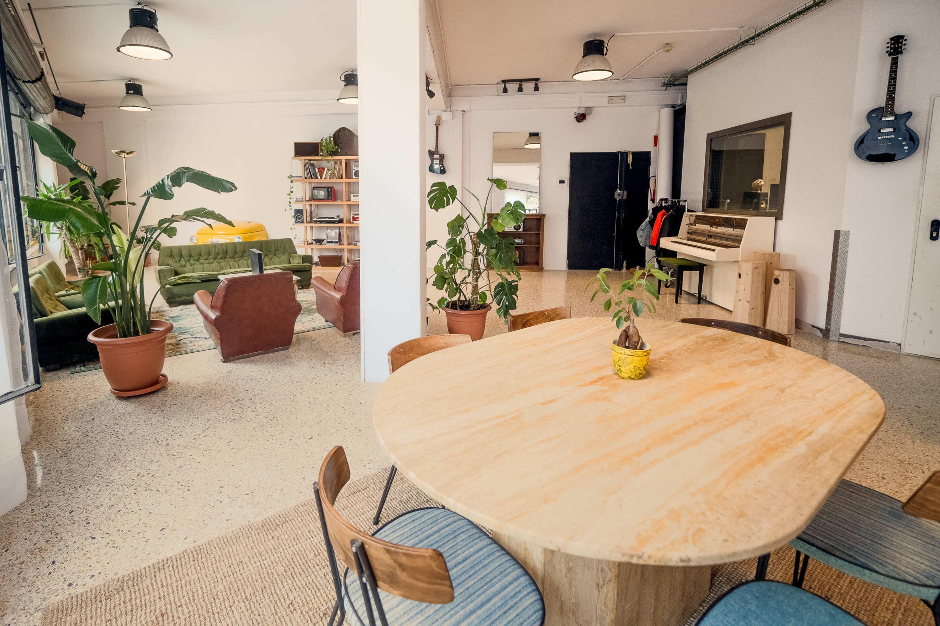 An interior space featuring a wooden dining table surrounded by chairs, with a living area that includes green and brown sofas, plants, and a musical instrument setup.