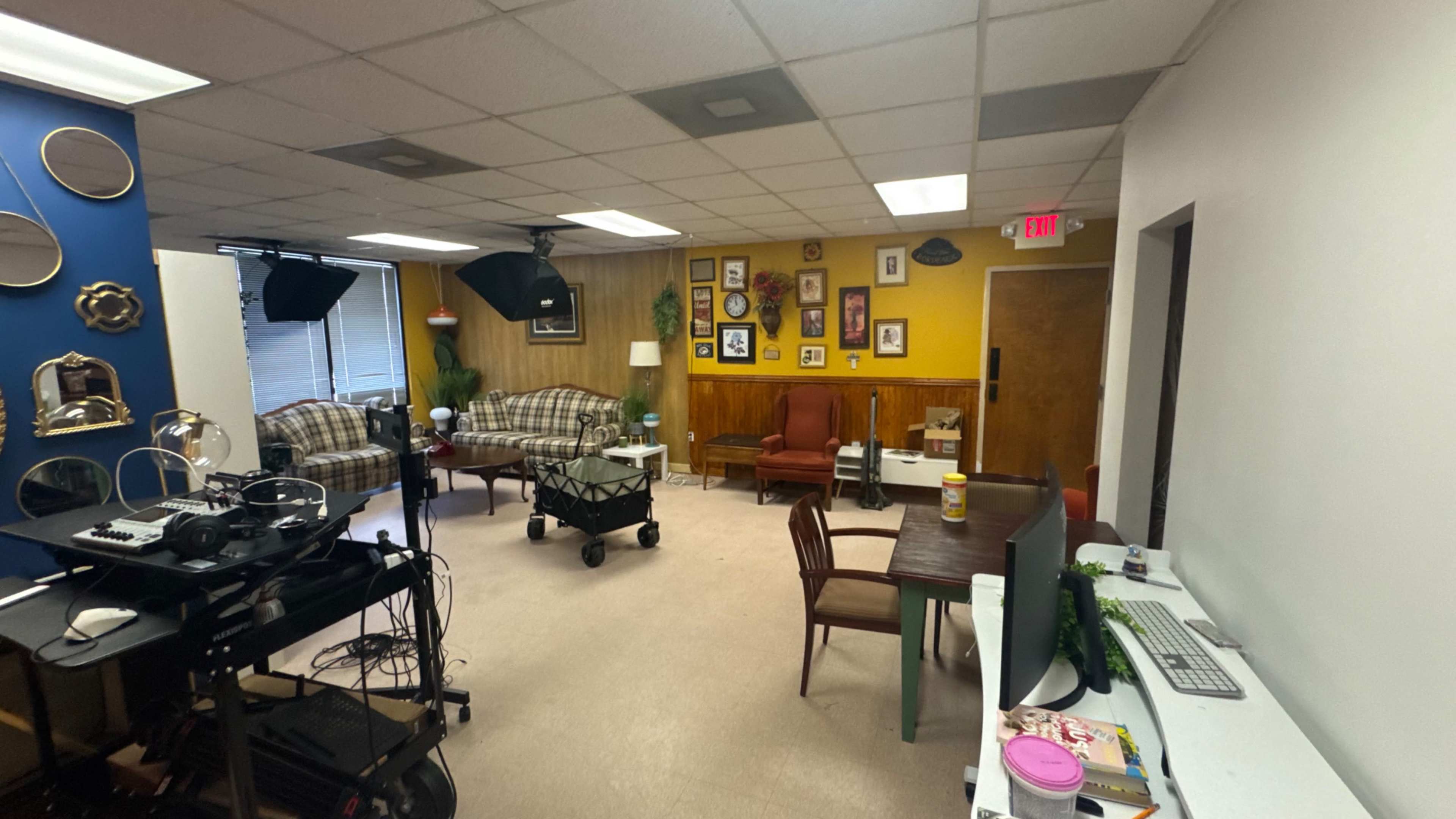 The image shows a spacious room with a mix of office and lounge furniture, including couches, chairs, a table, and a cart, along with various decorative items on the walls.