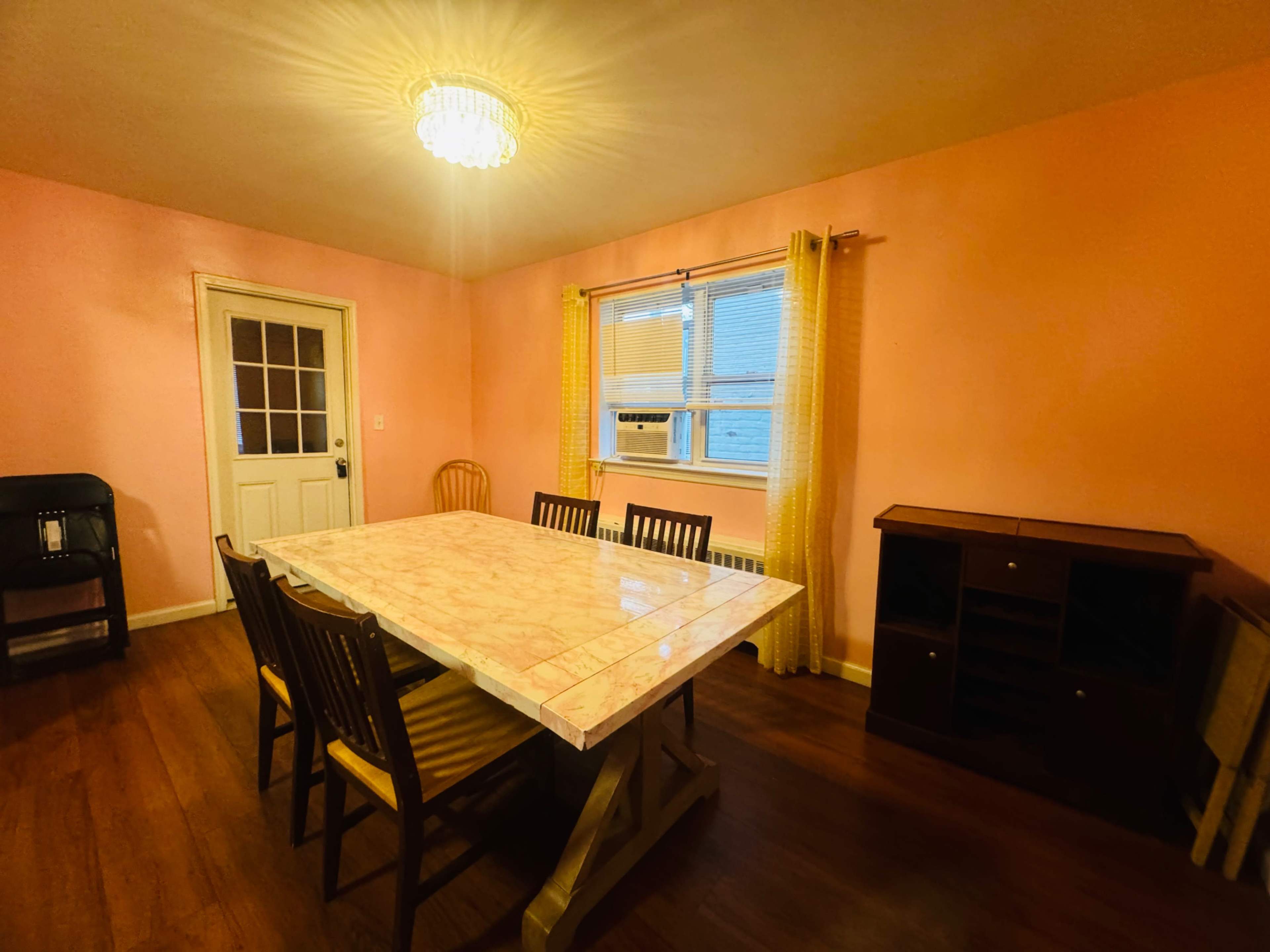 A dining room with a large table surrounded by wooden chairs, a door leading outside, and a window with yellow curtains, all set against pink walls.