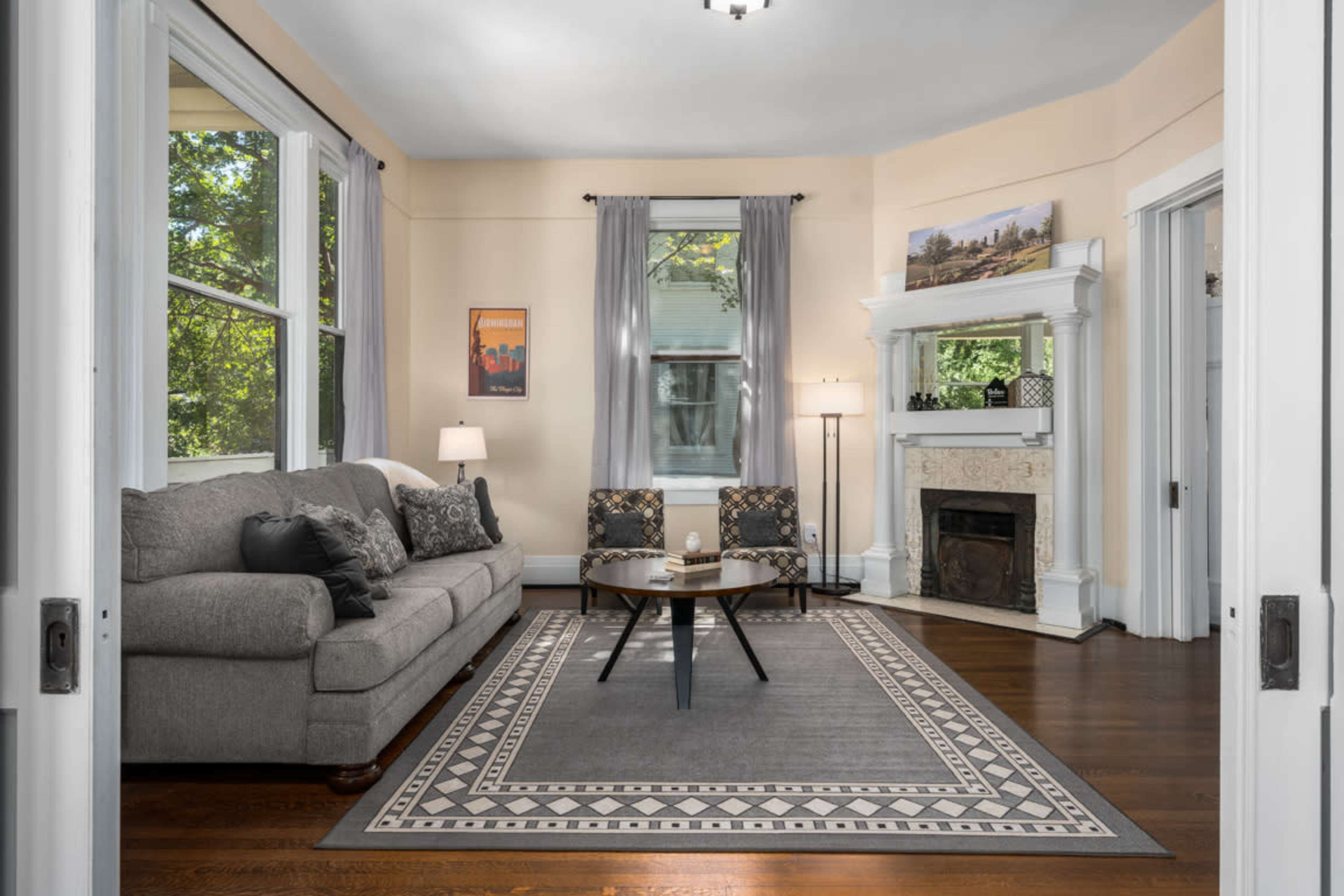 A living room features a gray sofa, a round coffee table, and a decorative fireplace, all framed by large windows letting in natural light.