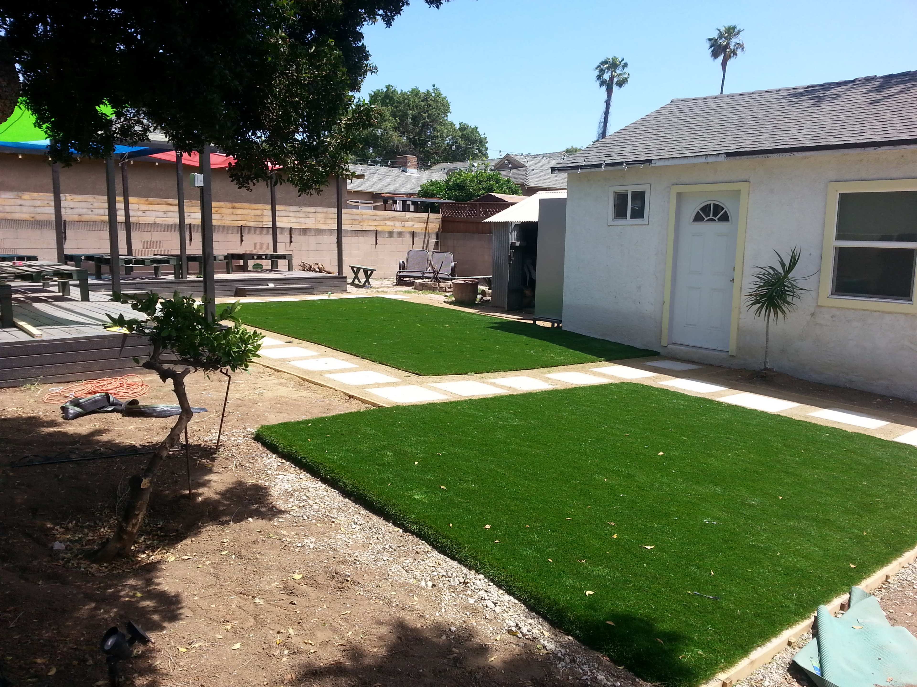 The image shows a landscaped area featuring two patches of artificial grass bordered by stone pathways, with a small white building and several palm trees in the background.