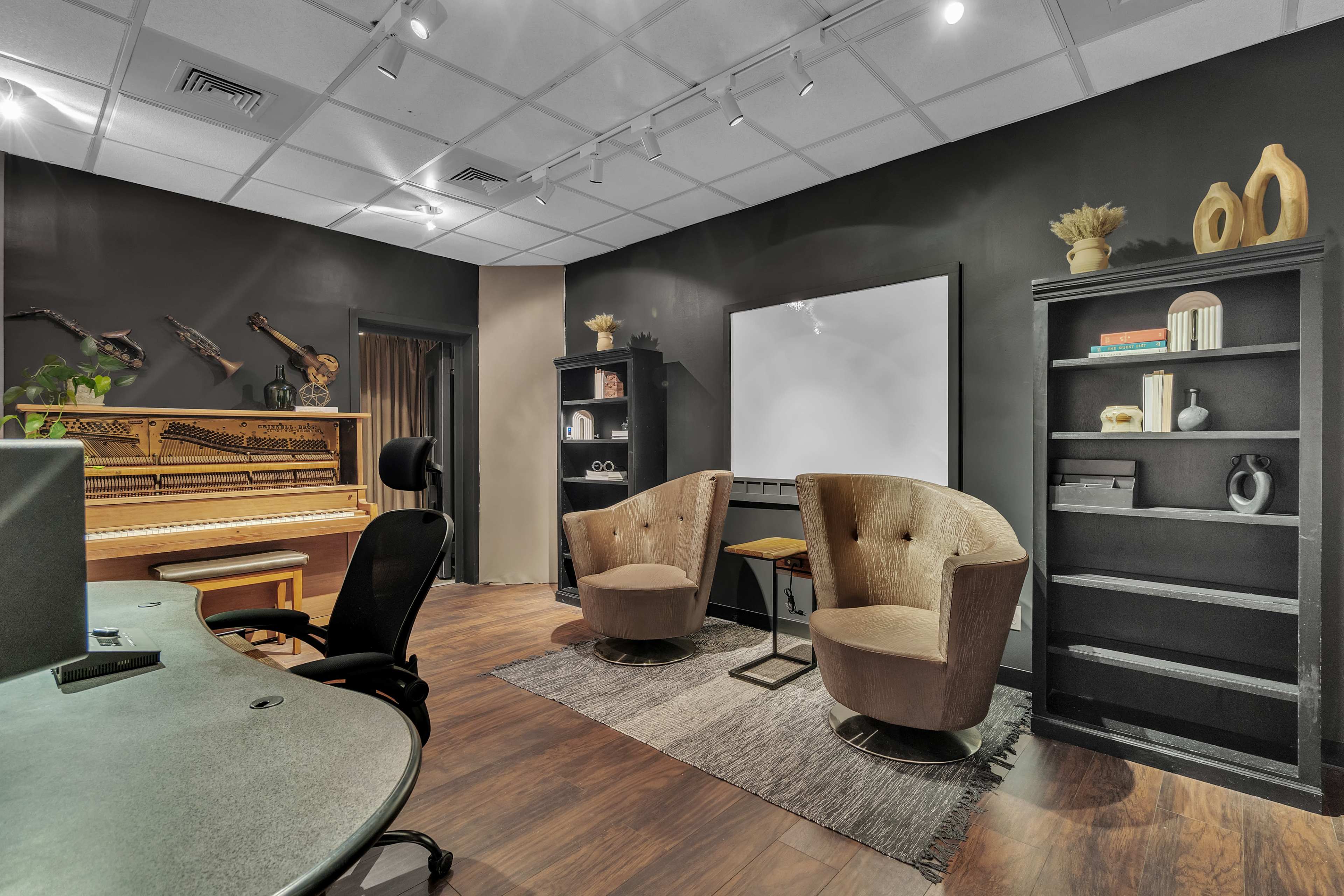 The image shows a modern room featuring a piano, two armchairs, a wall-mounted screen, and bookshelves with decorative items.