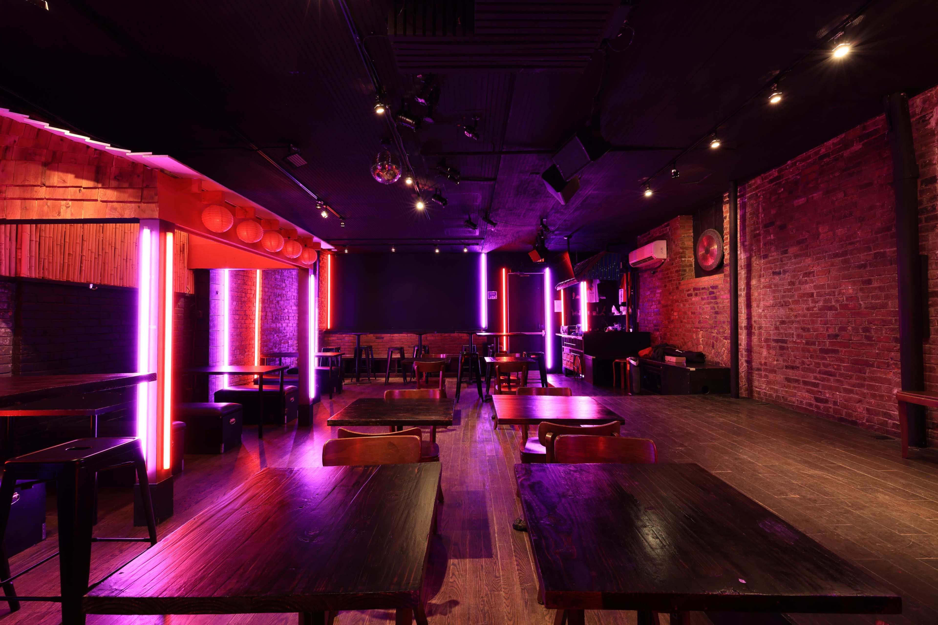 The image features a dimly lit, modern interior of a bar or lounge with wooden tables, exposed brick walls, and vibrant neon lighting.
