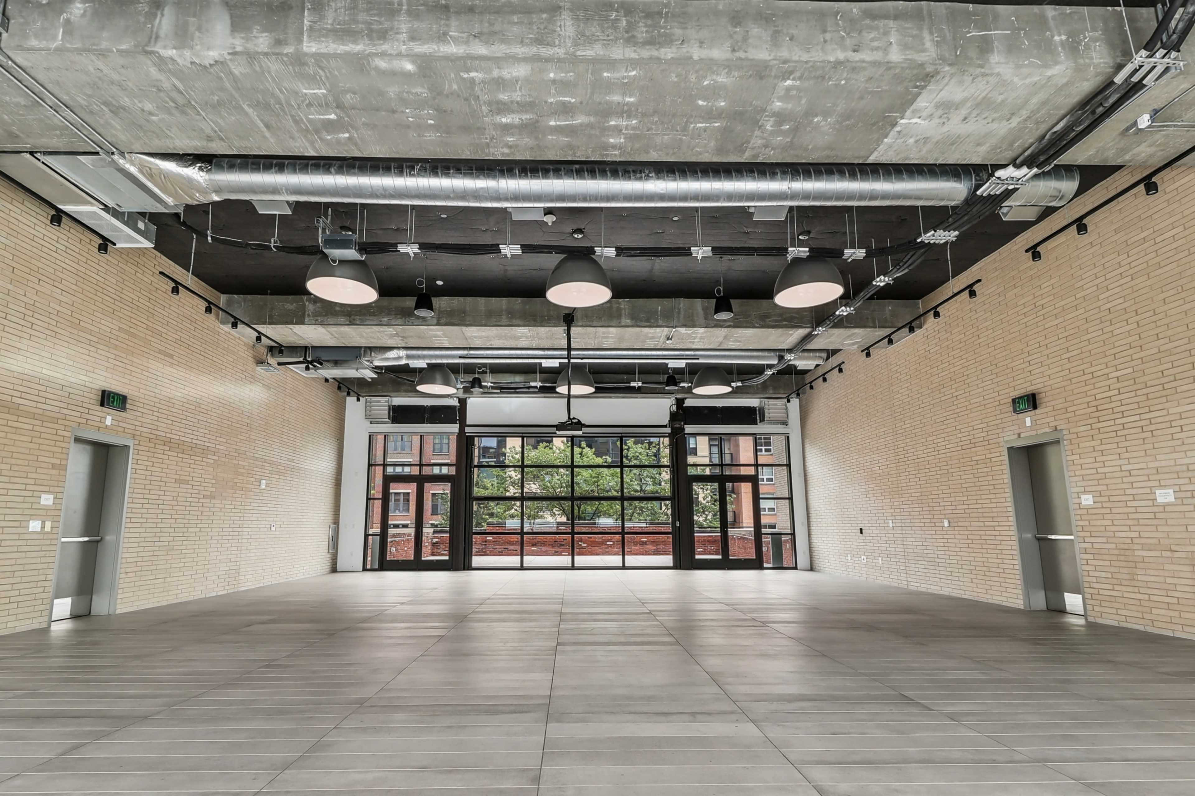 The image shows a spacious empty room with concrete walls, large windows, and industrial-style lighting fixtures.