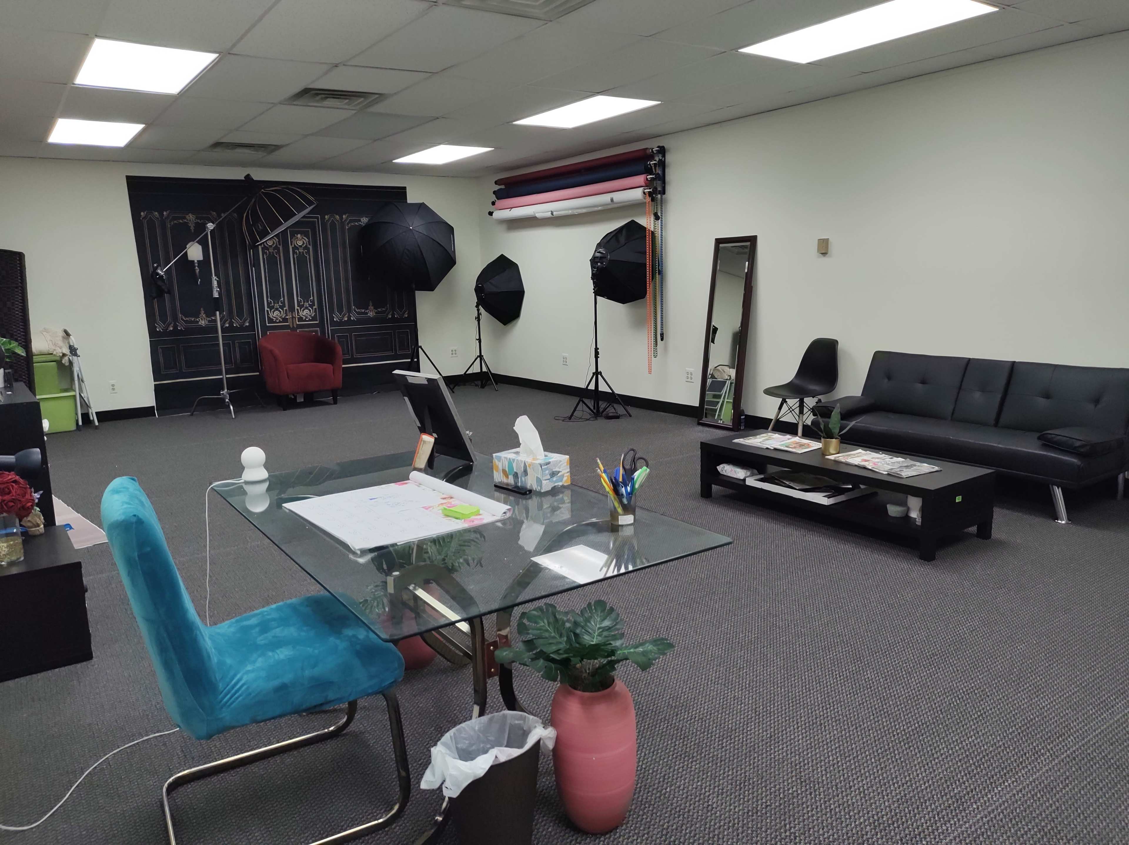 A spacious studio featuring a glass desk with a blue chair, a black sofa, a coffee table, and several photography lights positioned around the room.