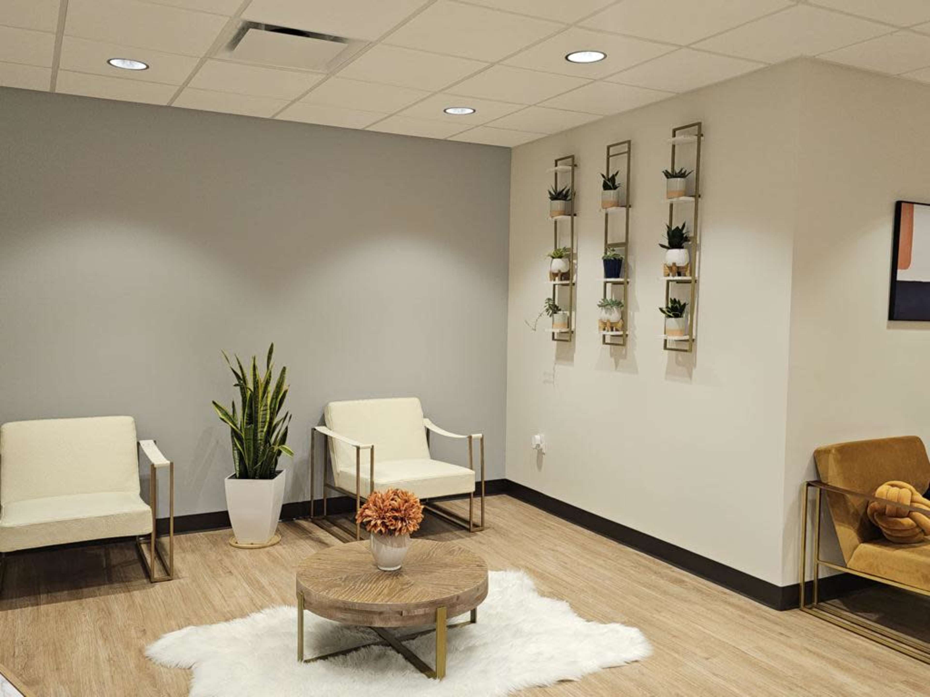 The image shows a modern waiting area with two beige armchairs, a round wooden coffee table on a white rug, and a wall-mounted planter displaying small plants.
