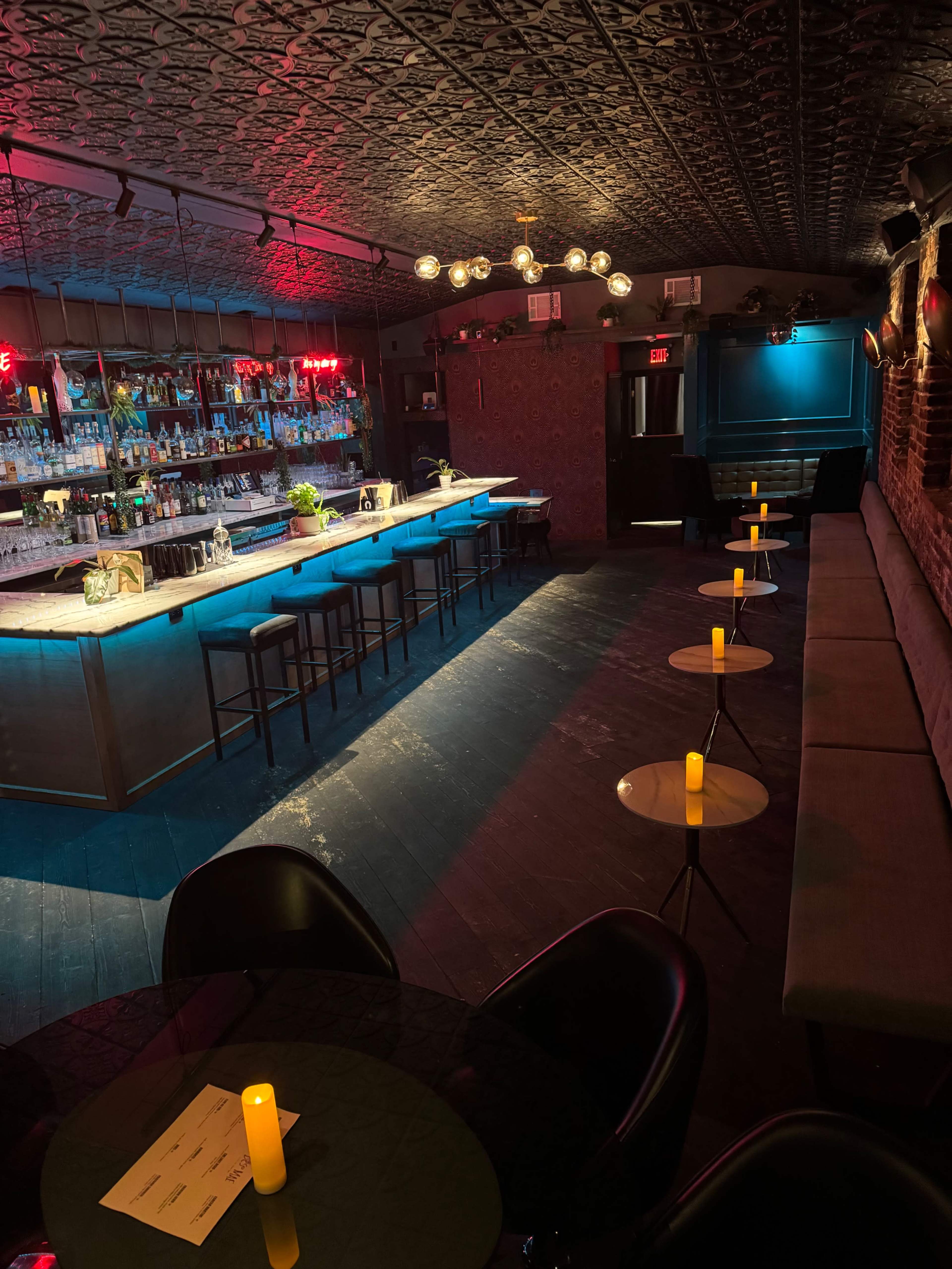 The image shows a dimly lit bar interior featuring a long counter with illuminated shelves of liquor, seating along the walls, and small tables with candles scattered throughout the space.