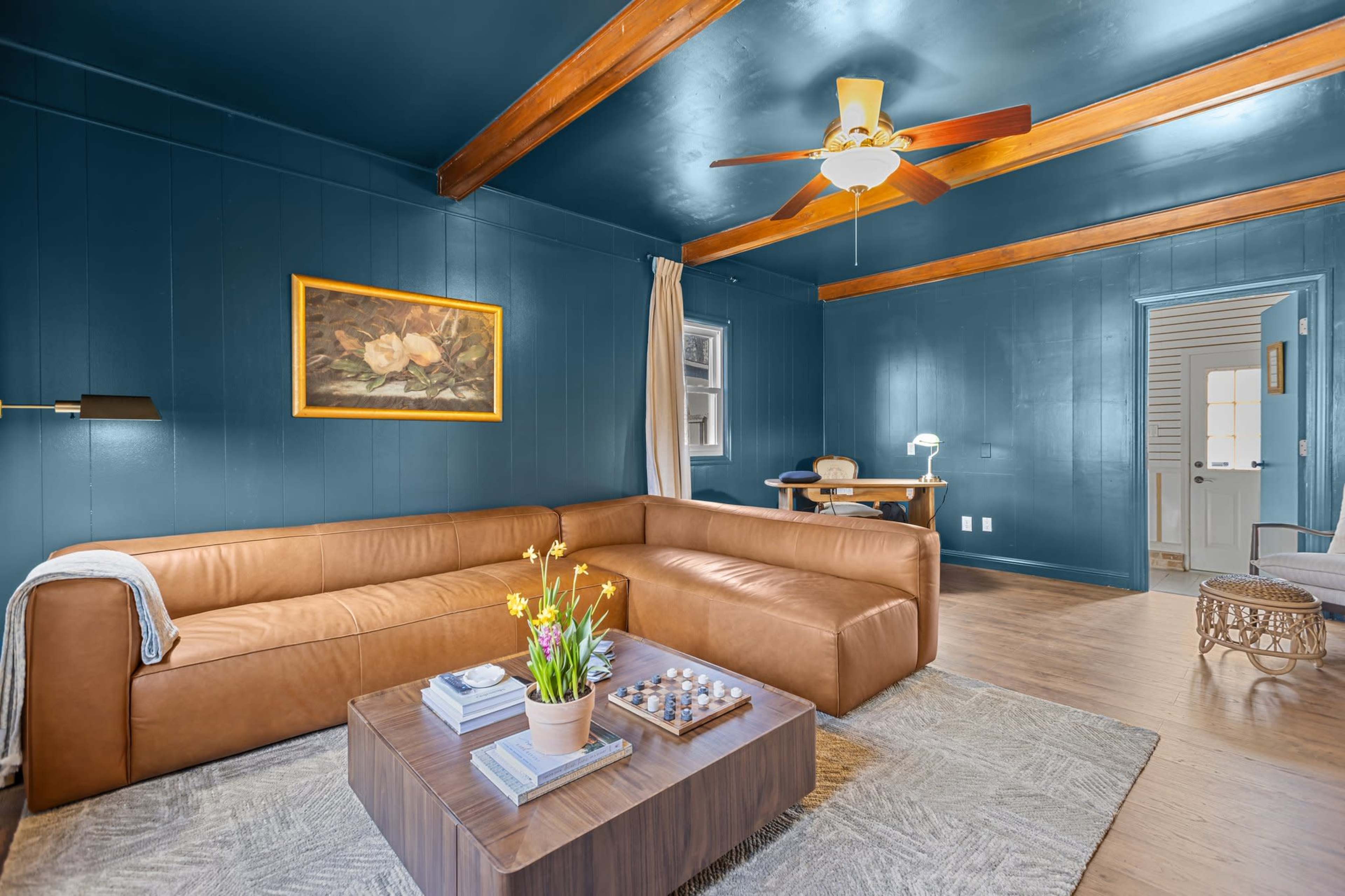 The image shows a modern living room with a leather sofa, coffee table with decorative items, and blue walls featuring wooden beams and a painting.