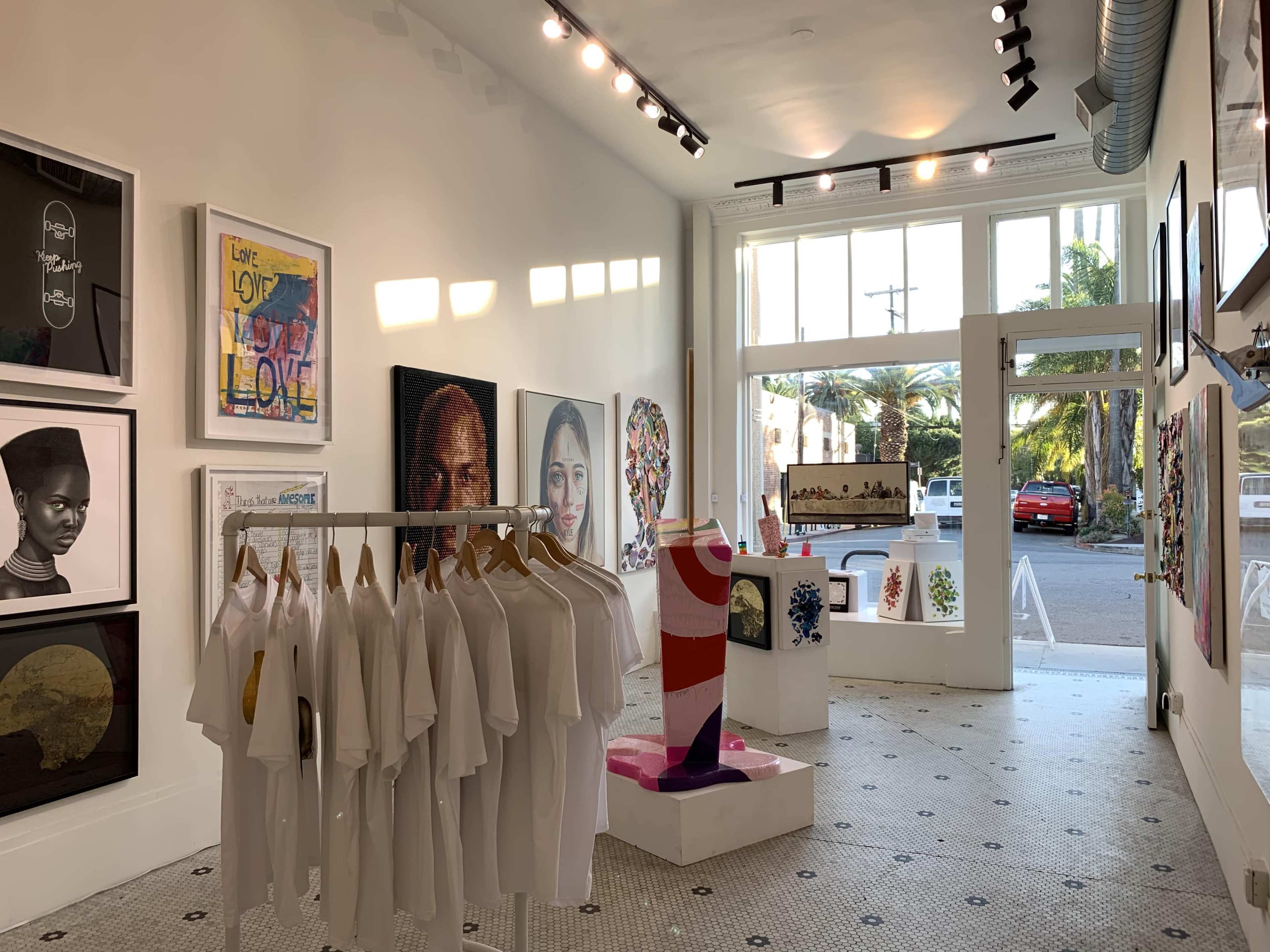 An art gallery featuring various framed artworks on the walls, a display of white t-shirts, and a colorful sculpture in the center.