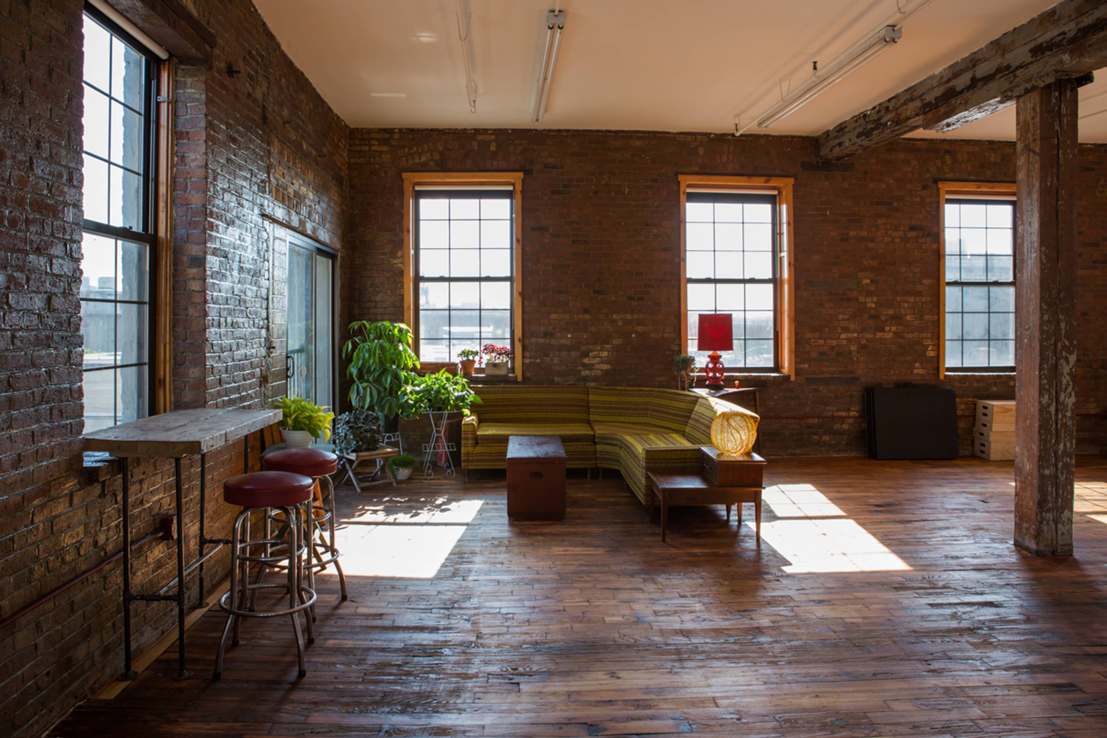 A spacious room with exposed brick walls, large windows, wooden floors, and a green couch accompanied by a wooden coffee table and bar stools.