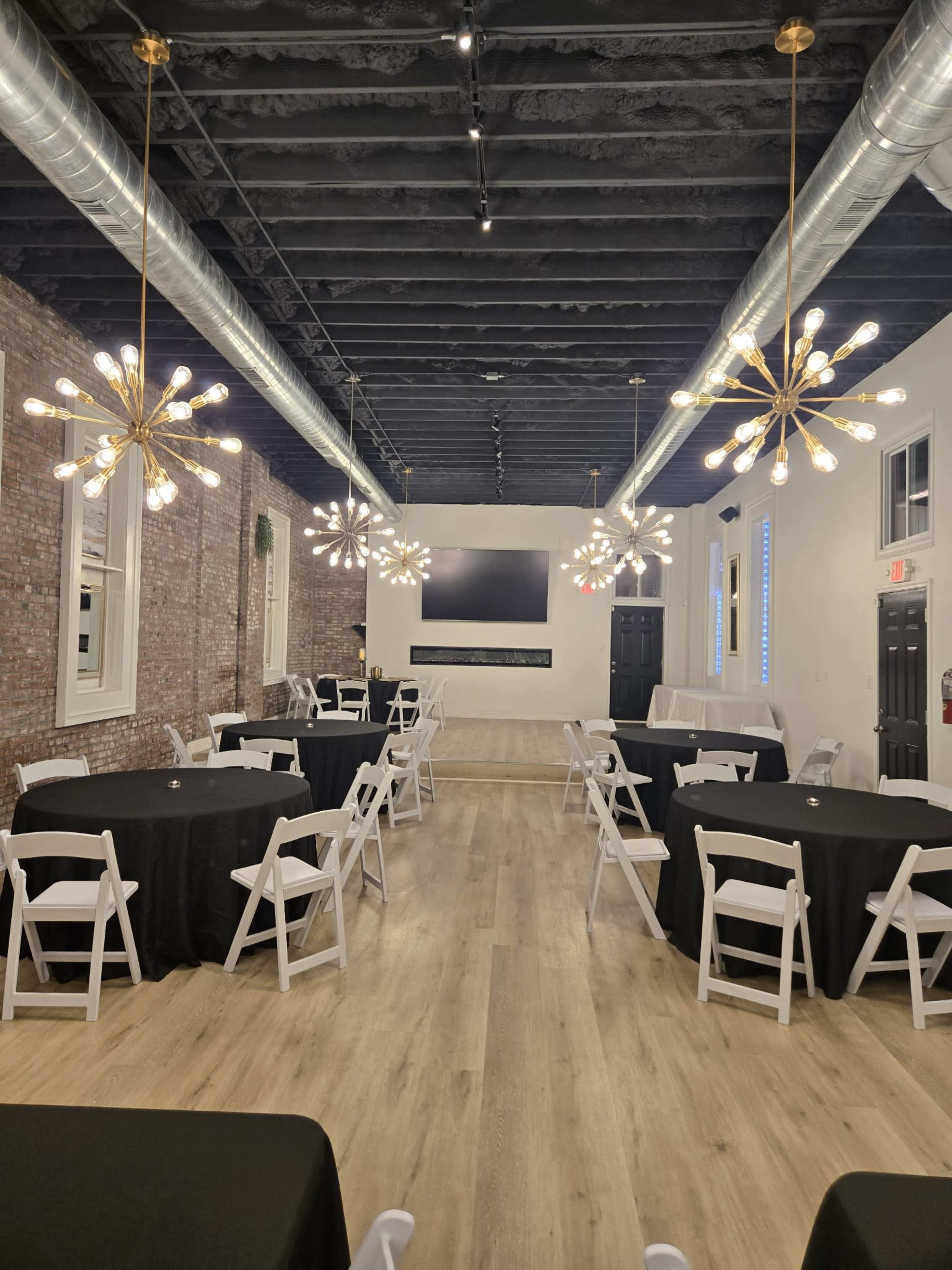 The image shows a spacious event venue with several round black tables and unique light fixtures hanging from the ceiling.