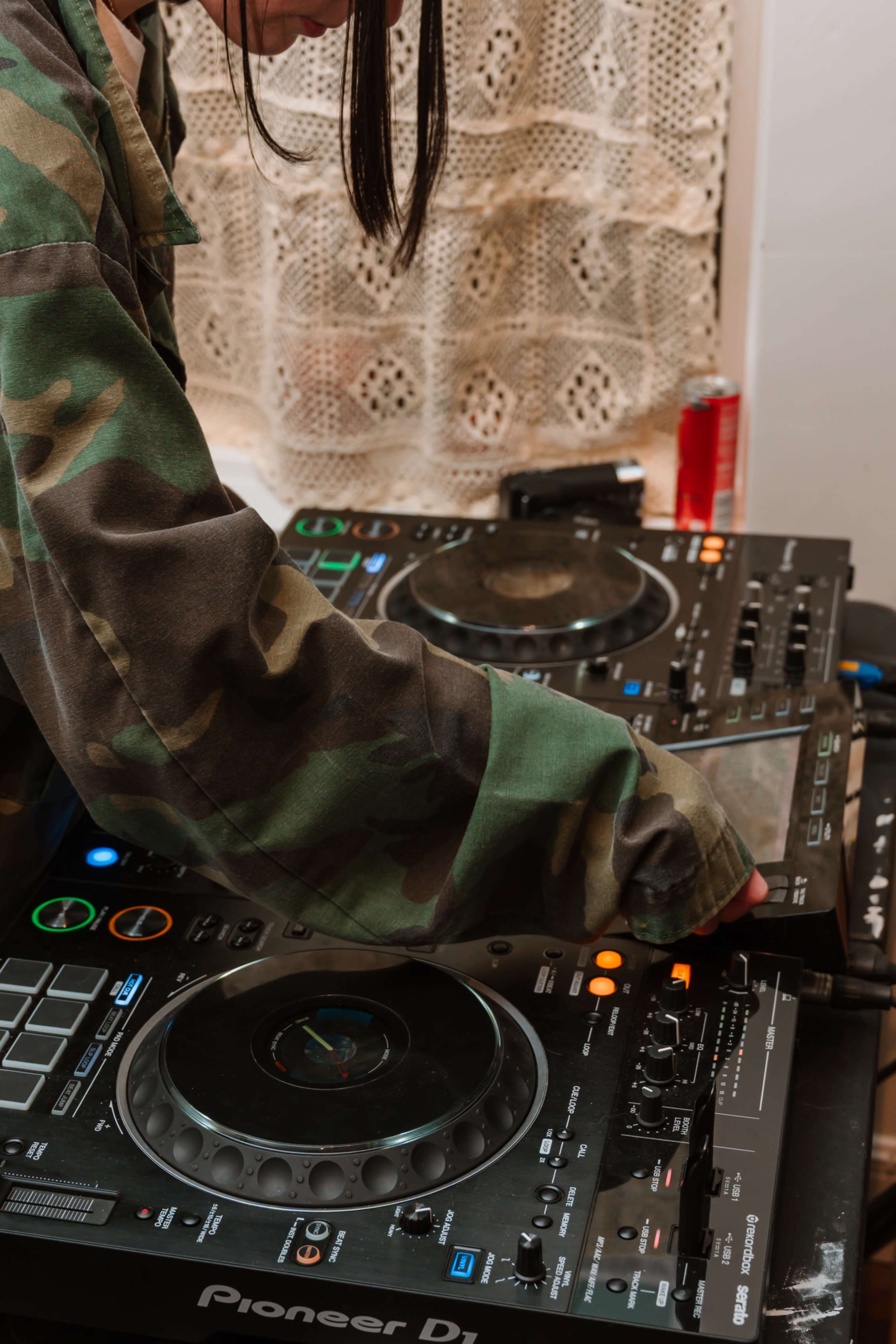 A person in a camouflage jacket adjusts the controls on a DJ mixer and turntables.