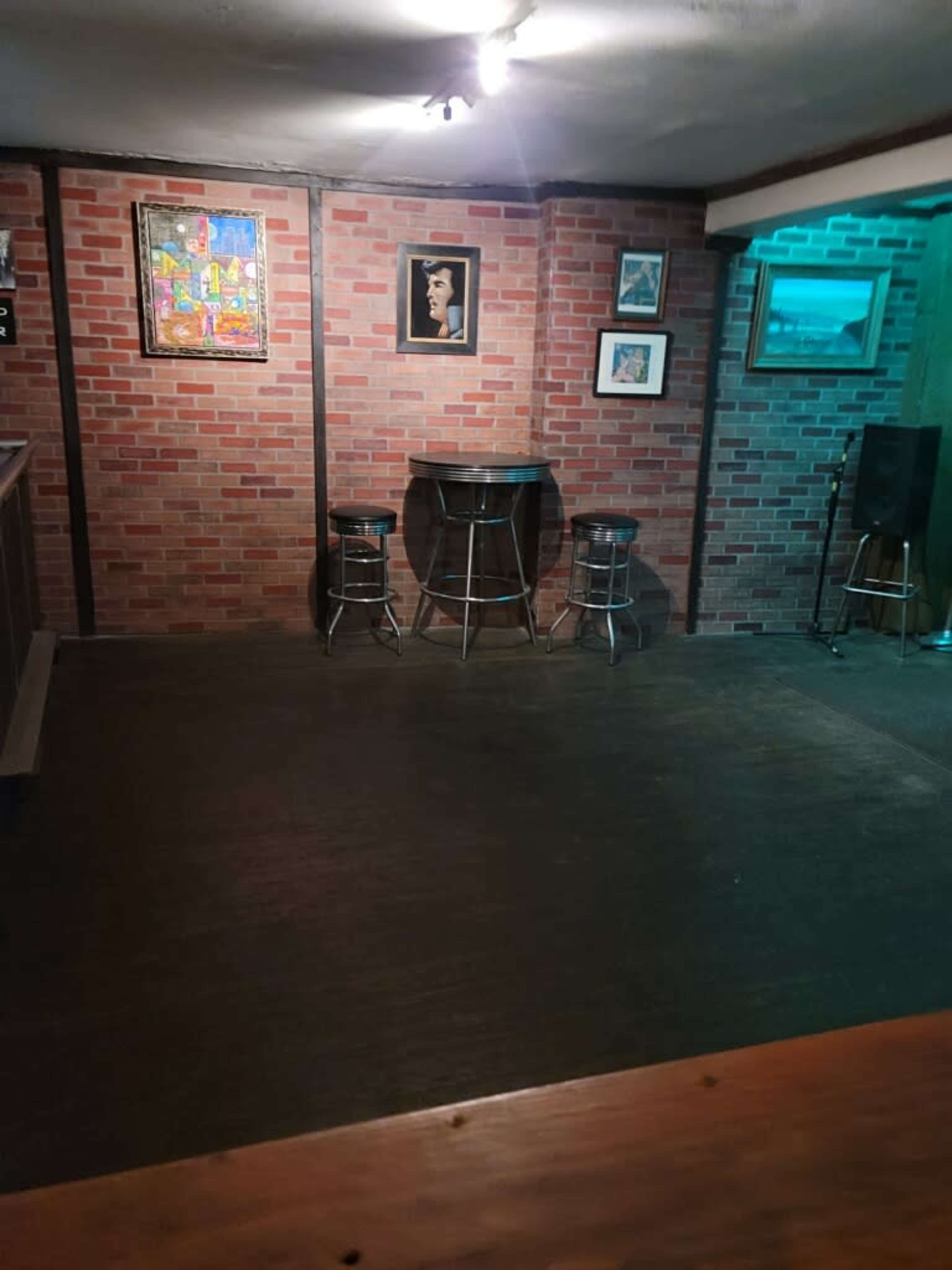 The image shows a small, dimly lit room with brick walls, a few bar stools, and various framed artworks.