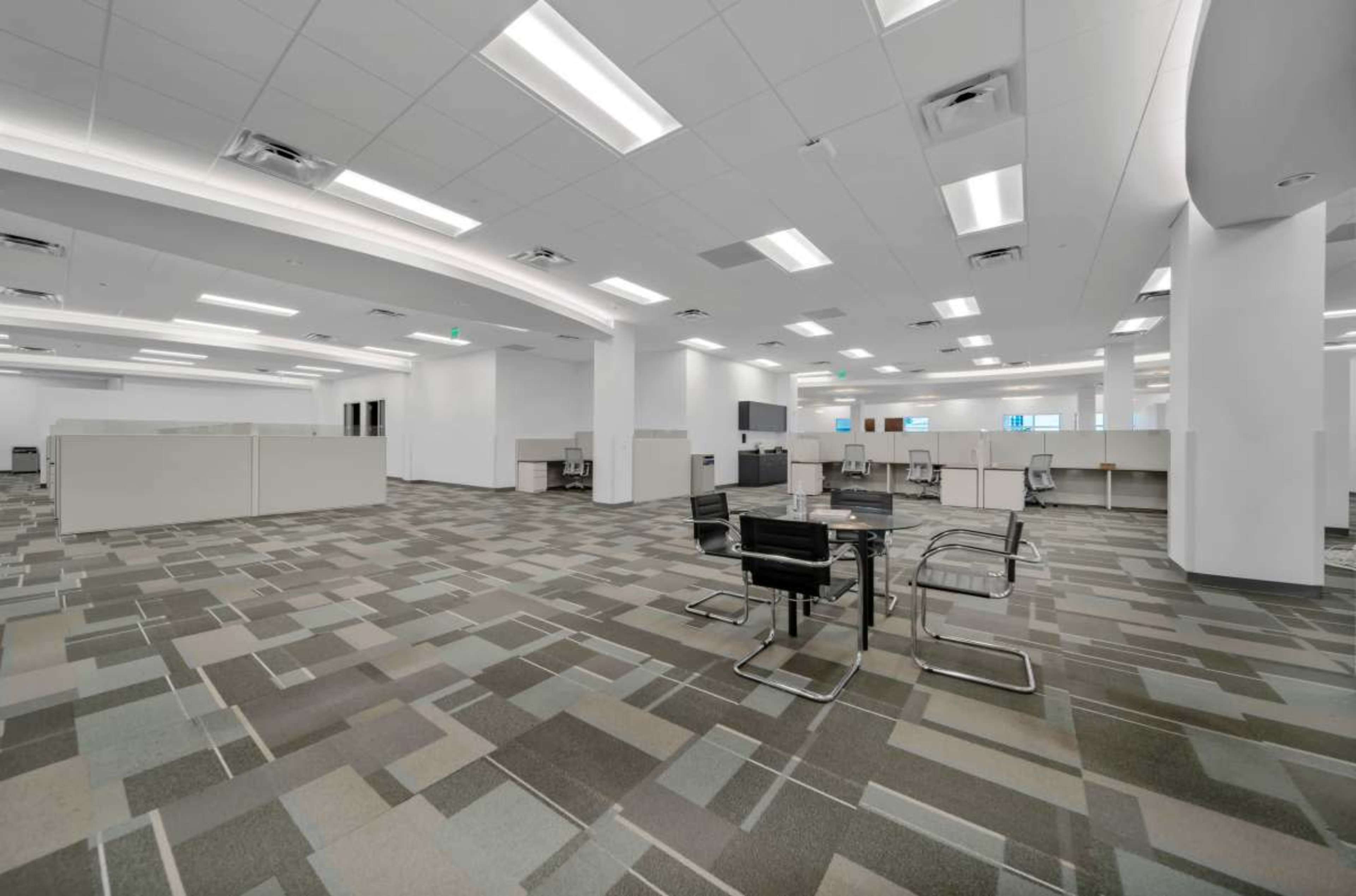 An empty office space features cubicles and a central seating area with a table and chairs on a patterned carpet.