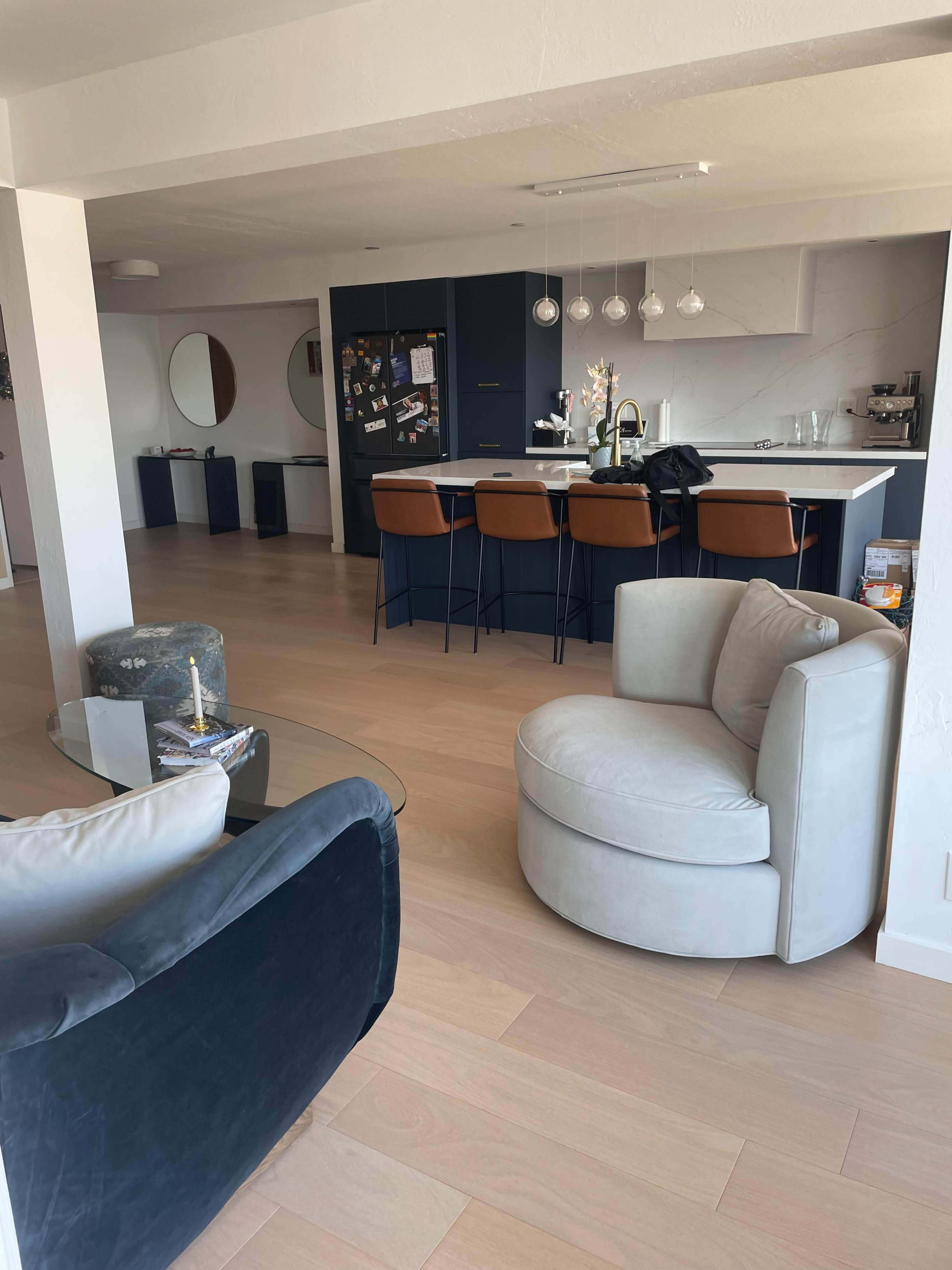 A modern open-concept living area with a kitchen featuring a dark-colored island, bar stools, and two distinct seating areas, including a round light-colored chair and a dark couch.