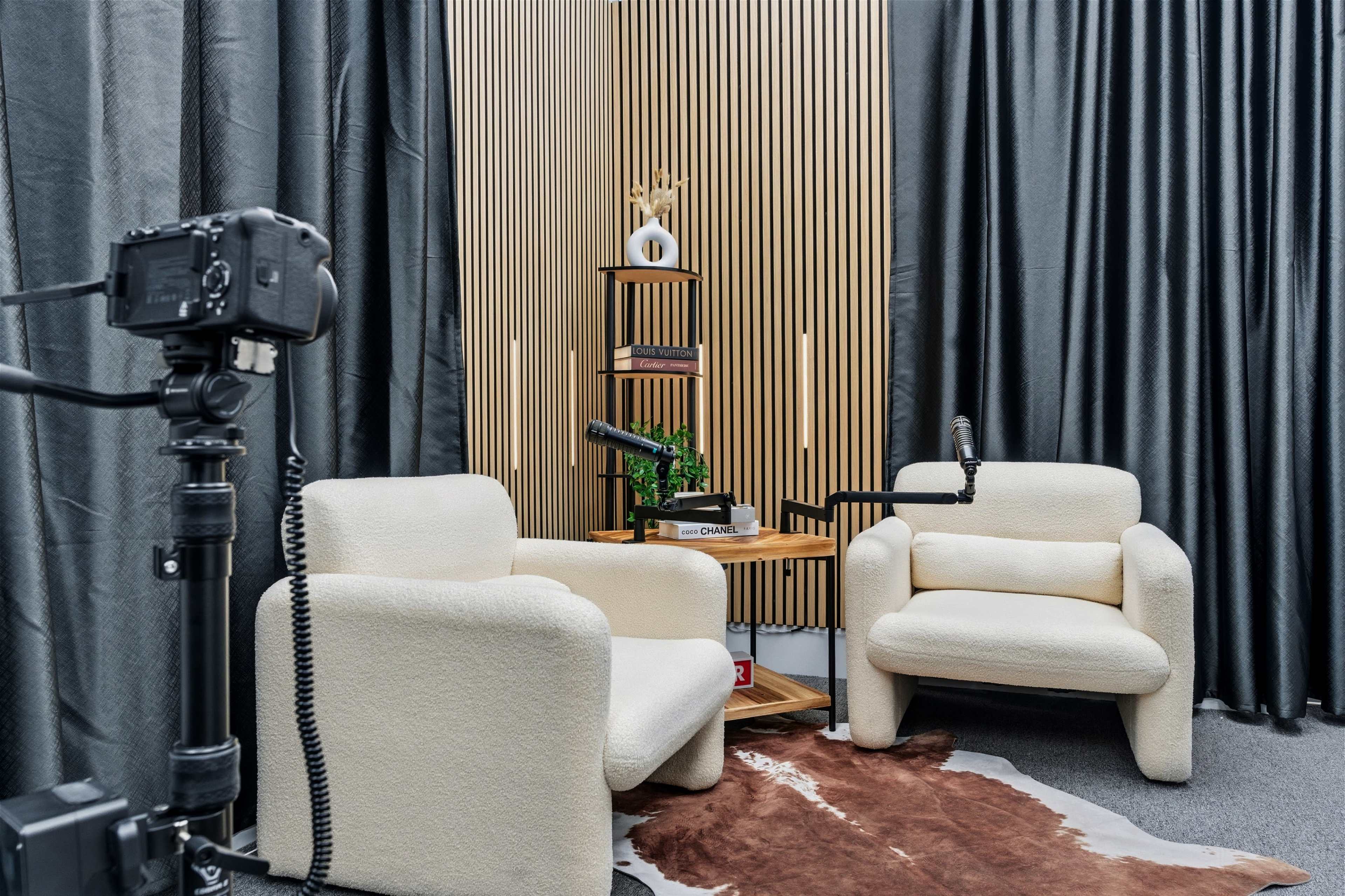 The image features a modern podcasting or recording studio setup with two plush armchairs positioned beside a wooden shelf, accented by a plant and two microphones on stands, all framed by dark curtains and a wooden panel backdrop.