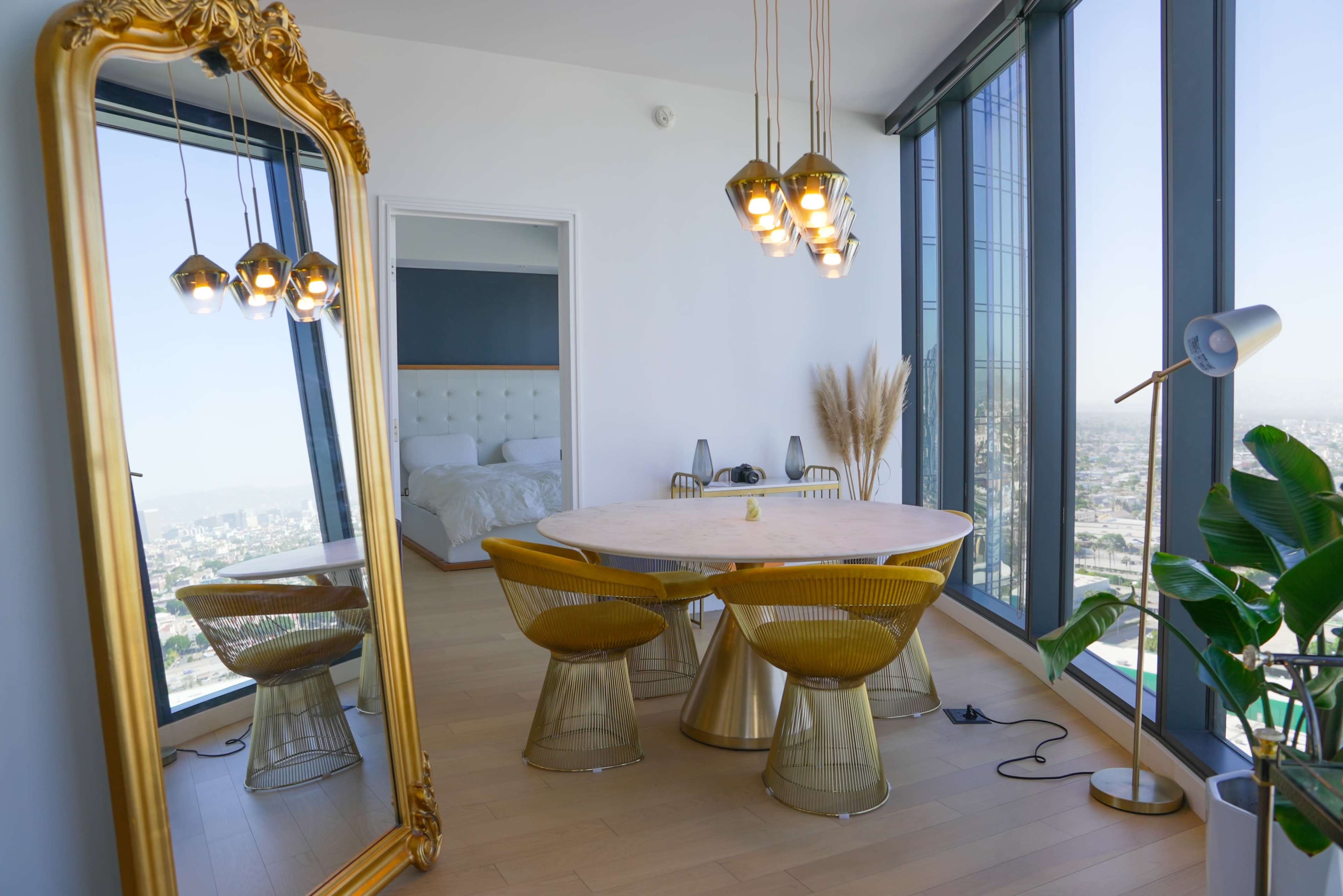 The image shows a modern dining area with a round marble table surrounded by four gold-accented chairs, a large mirror, and a floor lamp, all set against a backdrop of large windows and a city view.