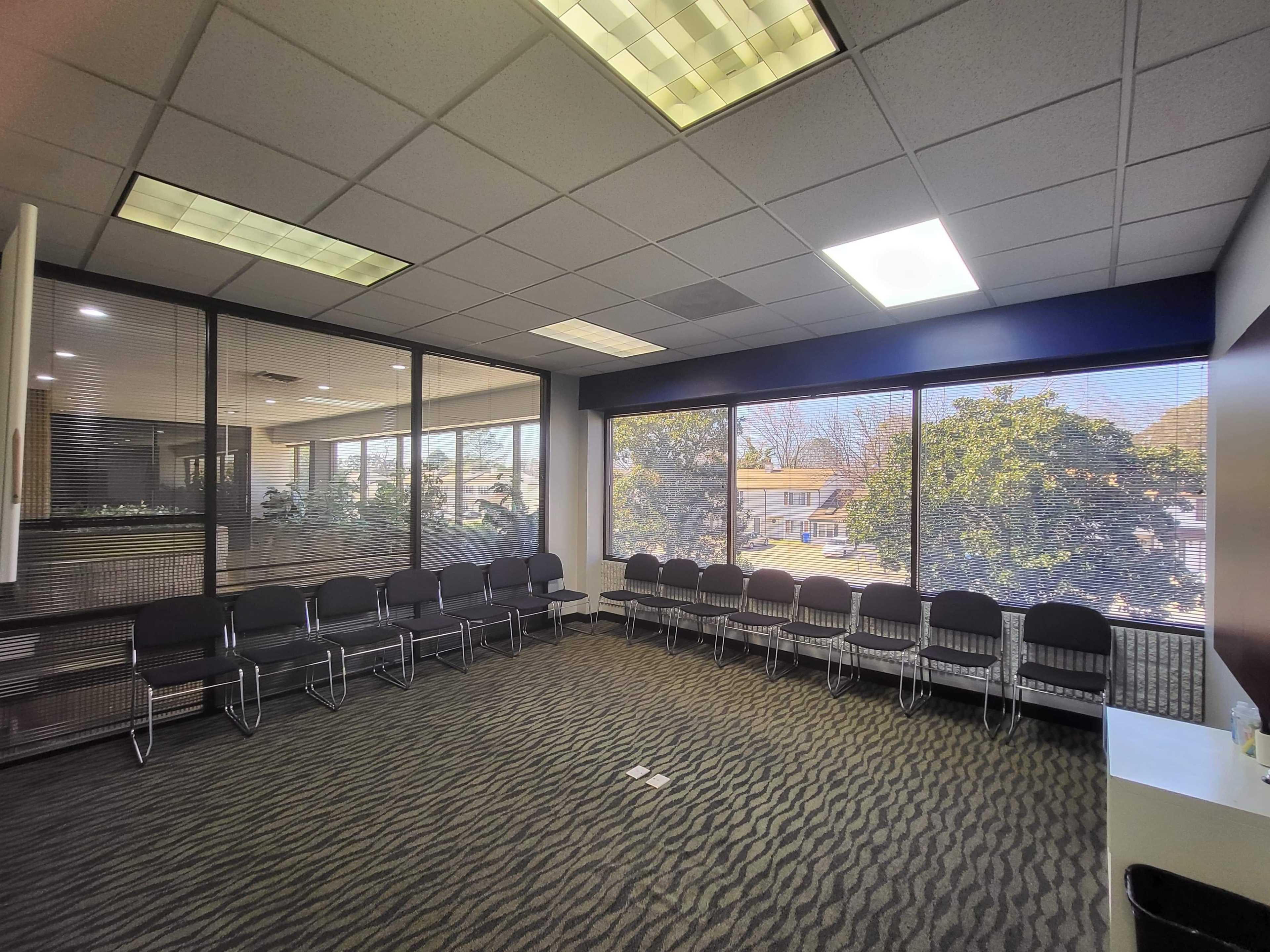 A waiting room features several chairs arranged in a circle, with large windows allowing natural light and a view of the outside.