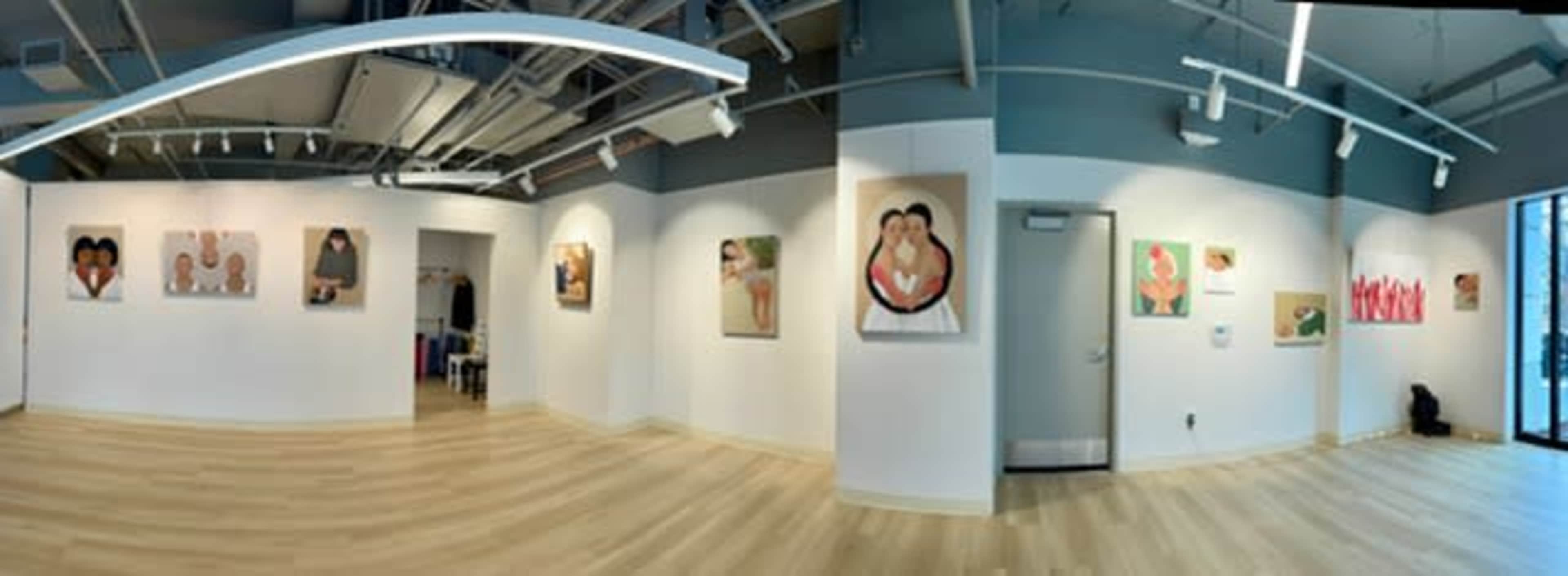 The image shows an art gallery interior with several paintings displayed on the white walls and wooden flooring.