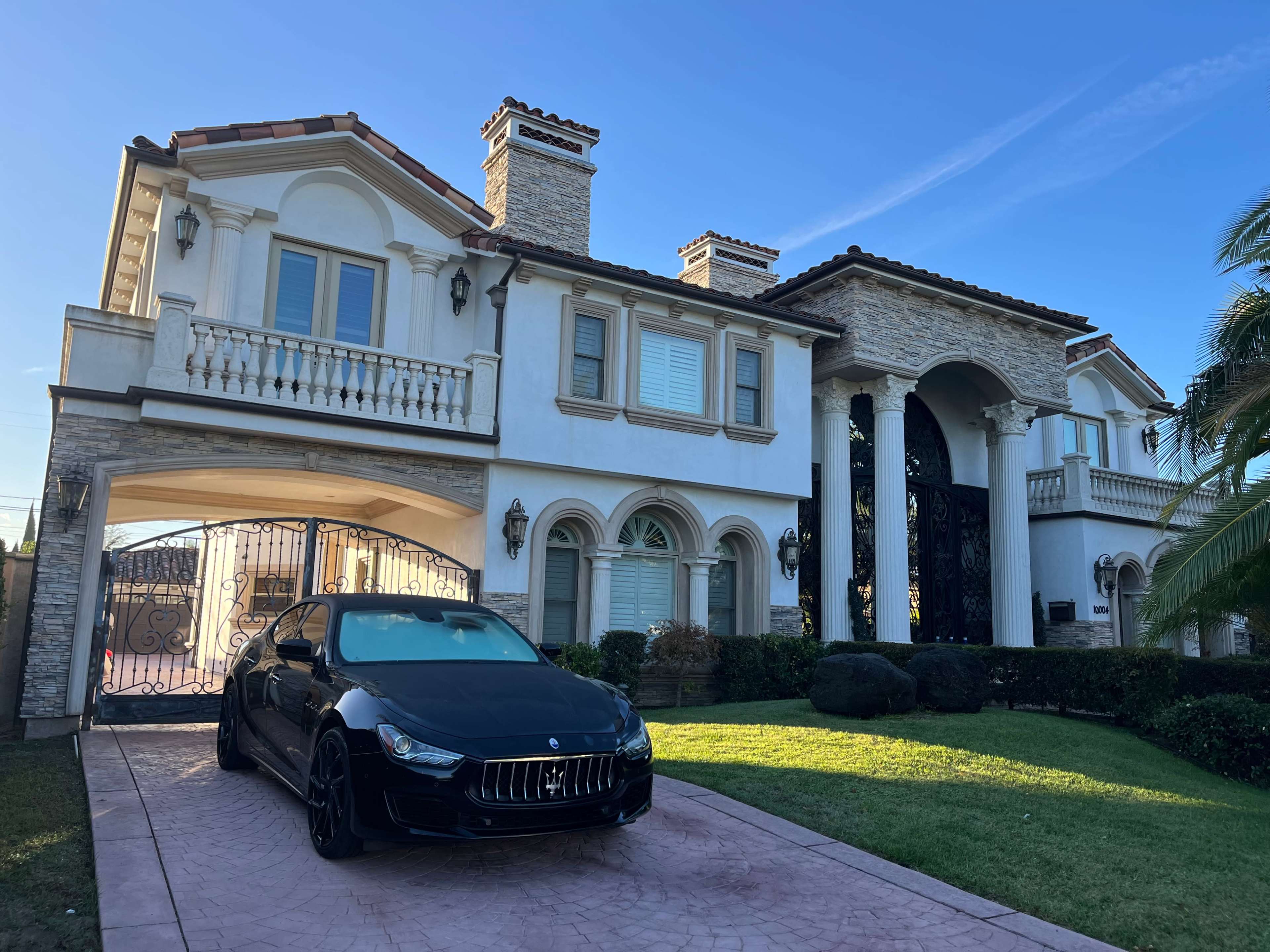 A large two-story house features a stone exterior, balconies, and a black car parked in the driveway.