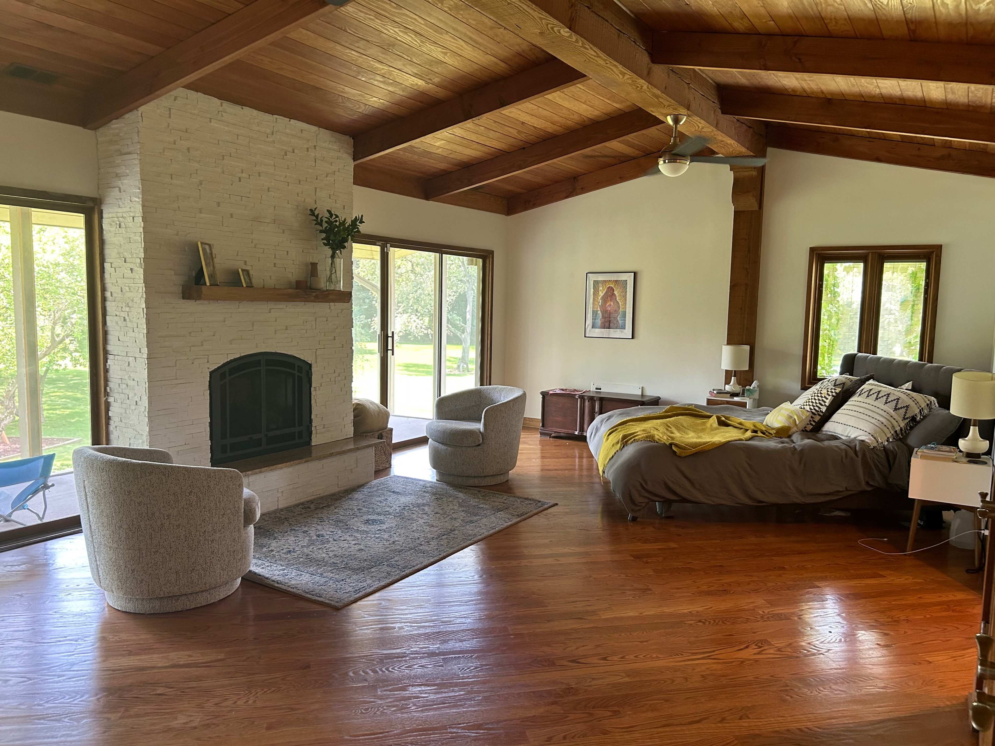 A spacious bedroom featuring a large bed, two armchairs, a stone fireplace, and windows that overlook a lush outdoor area.