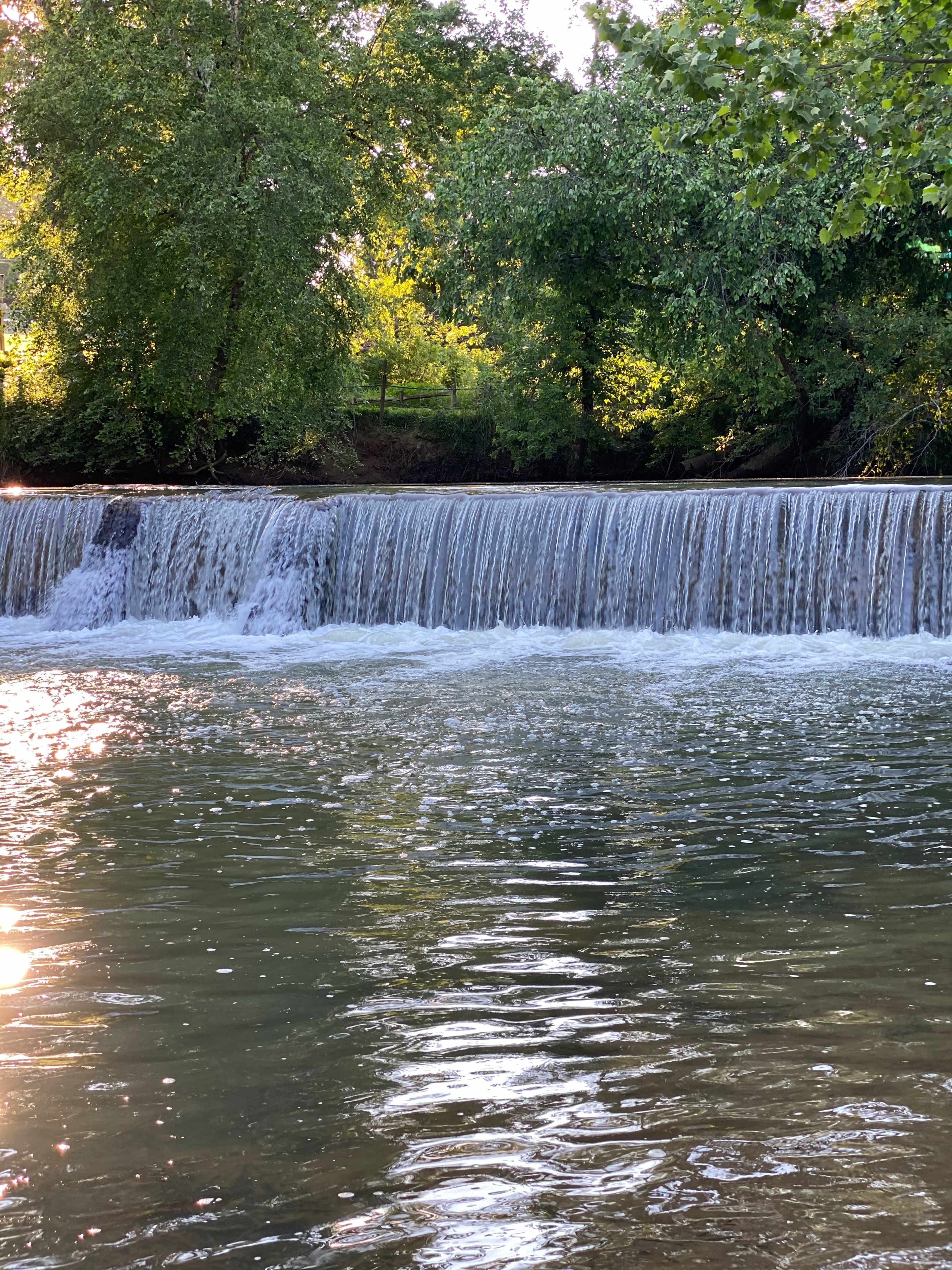 A waterfall cascades over a stone ledge into a serene pool, surrounded by lush greenery.