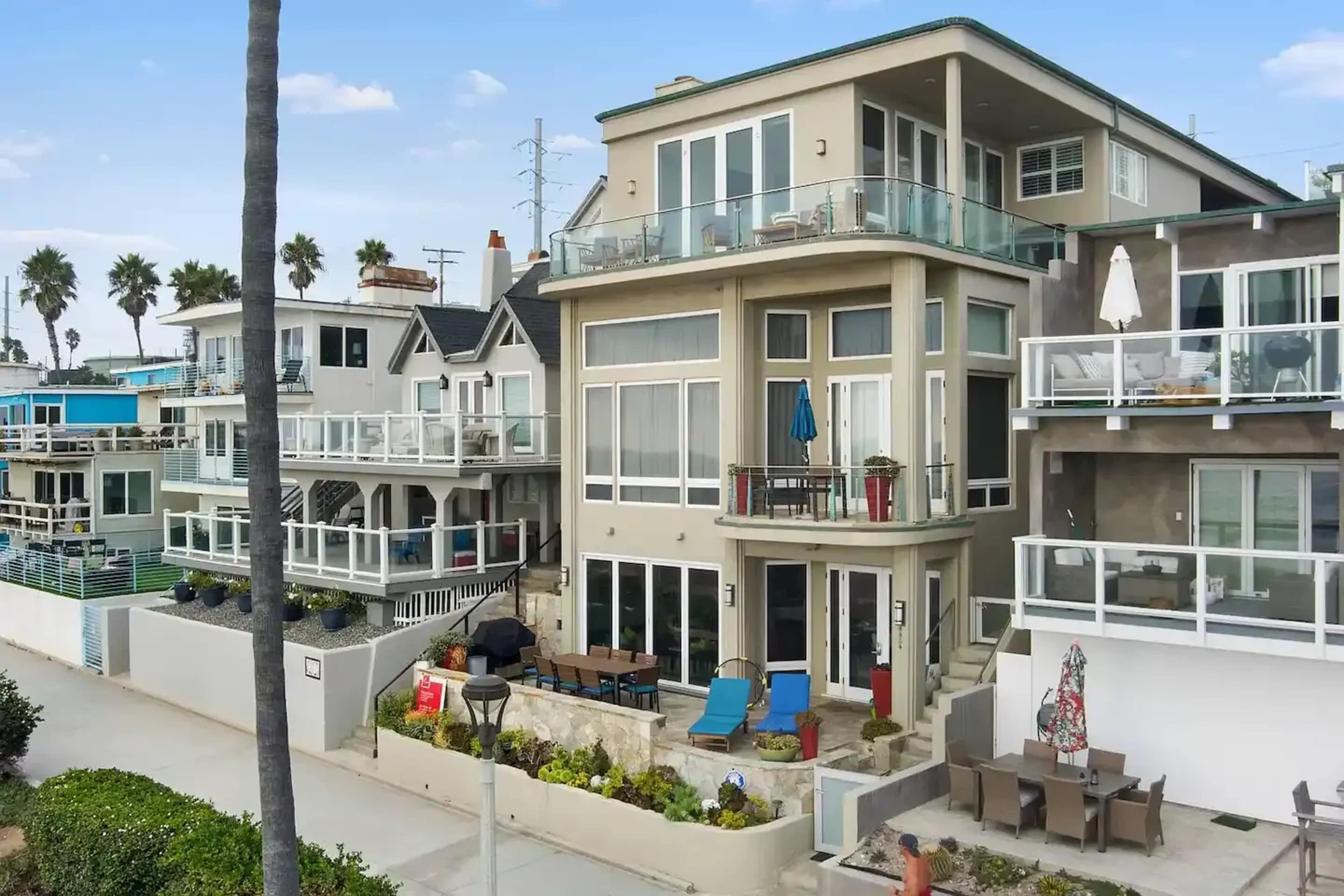 A modern beachfront house with large windows and a balcony, surrounded by neighboring houses and palm trees.