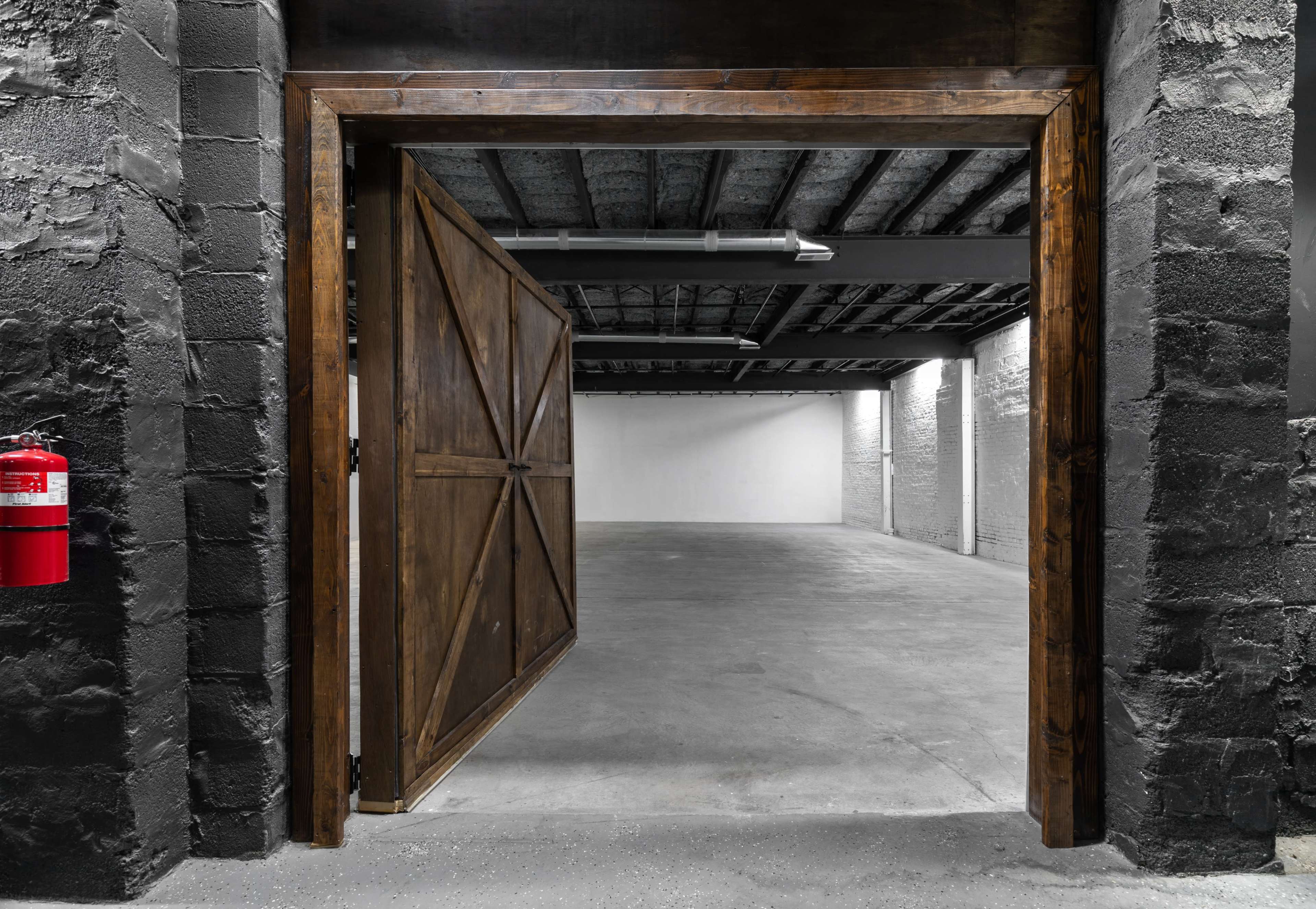 A wooden double door opens to reveal a spacious, empty room with concrete flooring and a partially exposed ceiling.