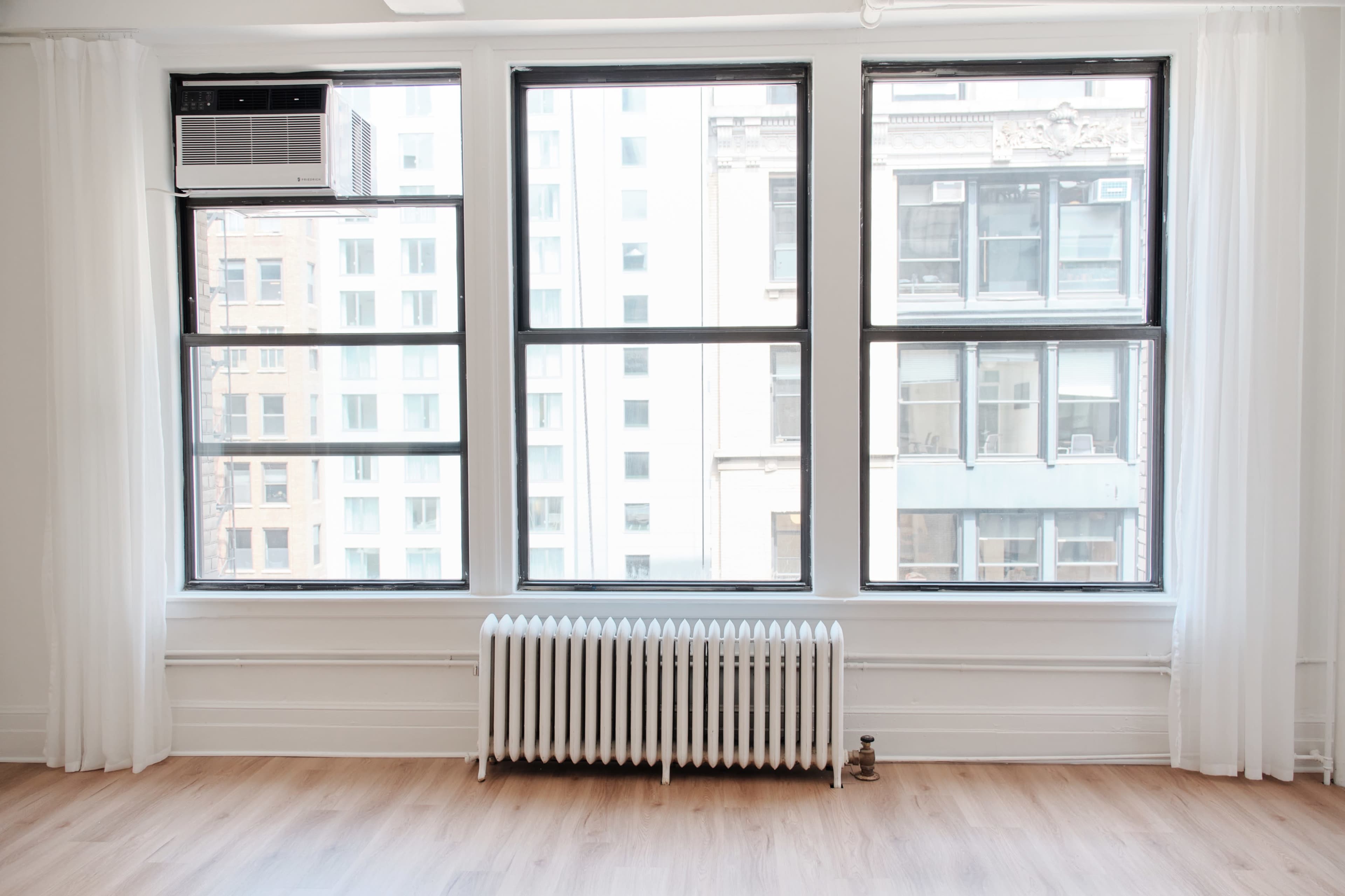 The image shows a room with three large windows overlooking a cityscape, featuring a radiator beneath the windows and sheer curtains on either side.