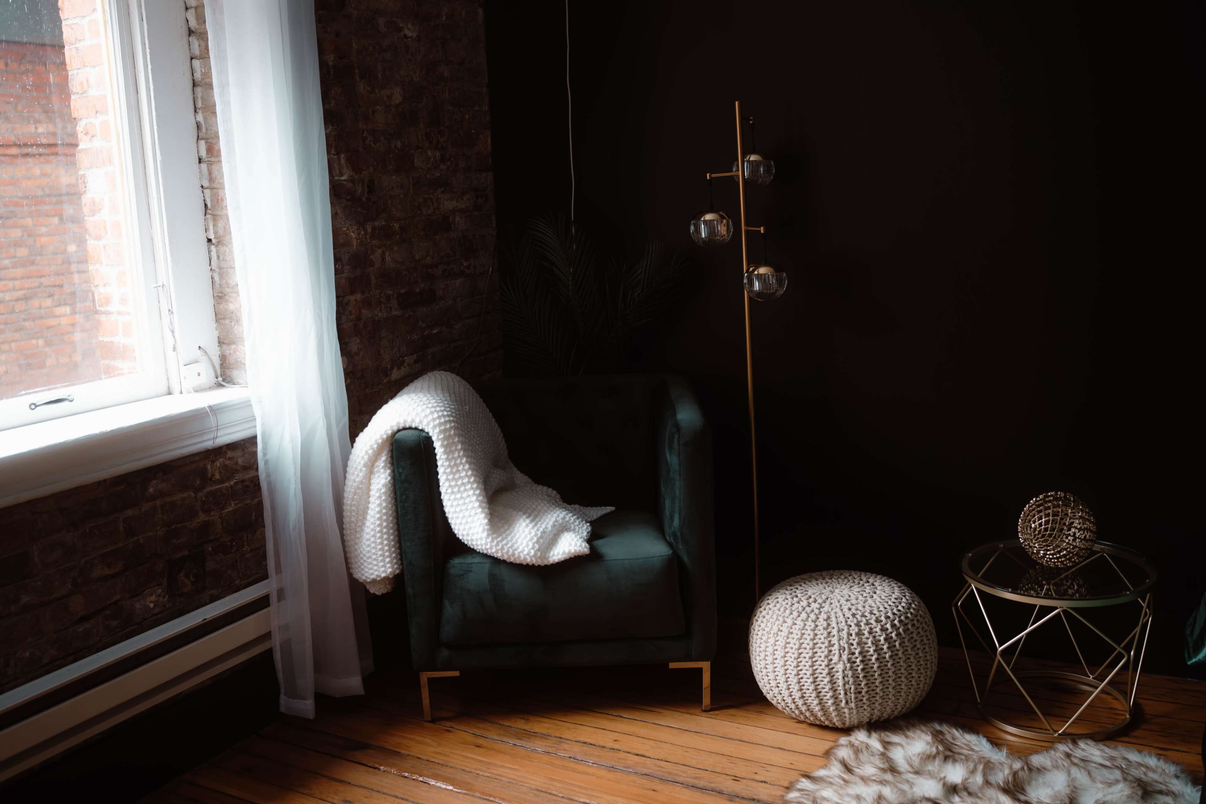 A green armchair with a white blanket draped over it is positioned beside a gold floor lamp and a round, knitted pouf in a room with exposed brick walls and wooden flooring.