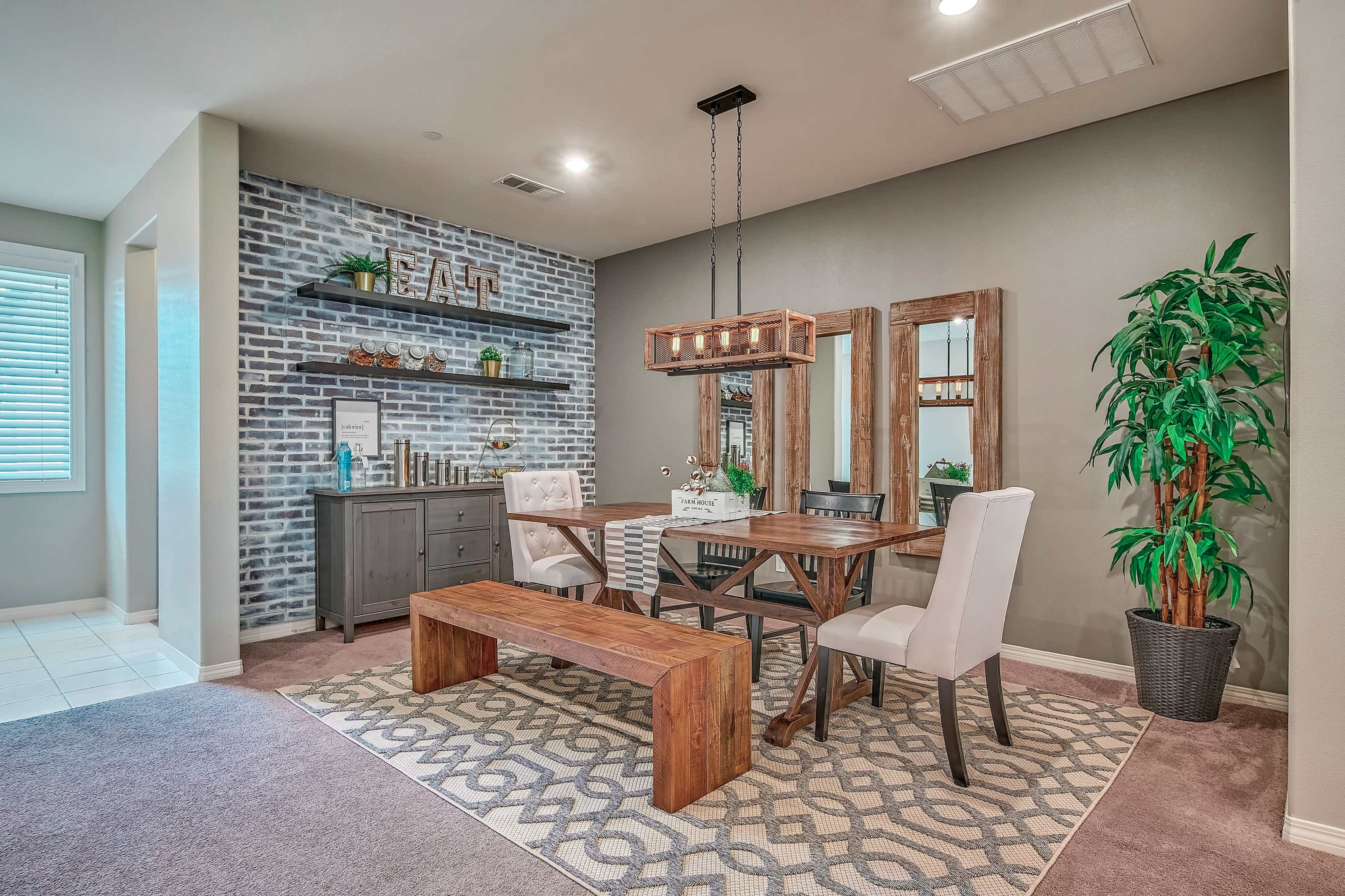 A dining area with a wooden table and chairs surrounded by a patterned rug, a gray sideboard, decorative mirrors, and a plant, against a backdrop of a brick feature wall.