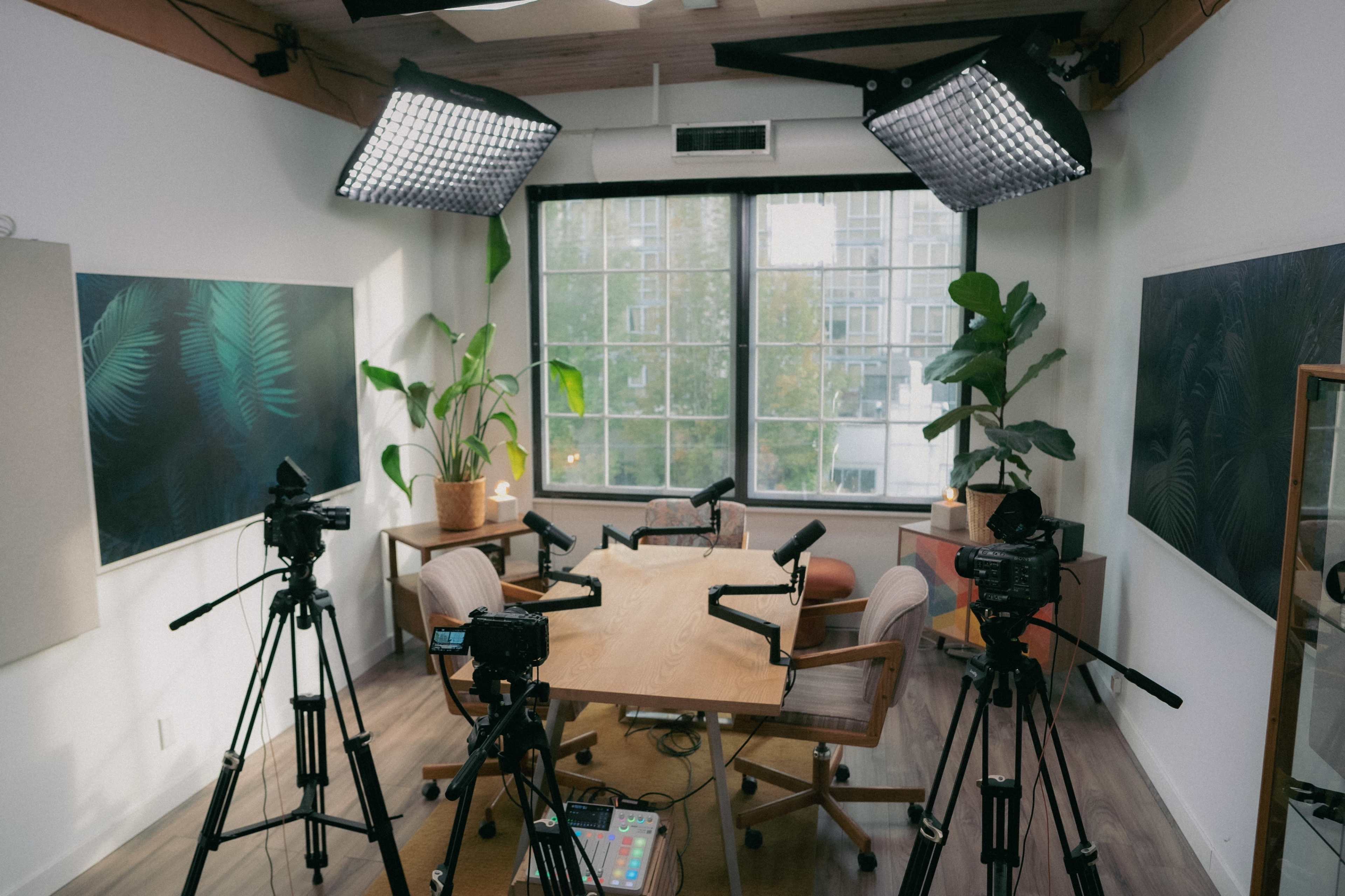 The image shows a well-lit recording studio featuring a wooden table surrounded by chairs, with several cameras set up and large windows providing natural light.