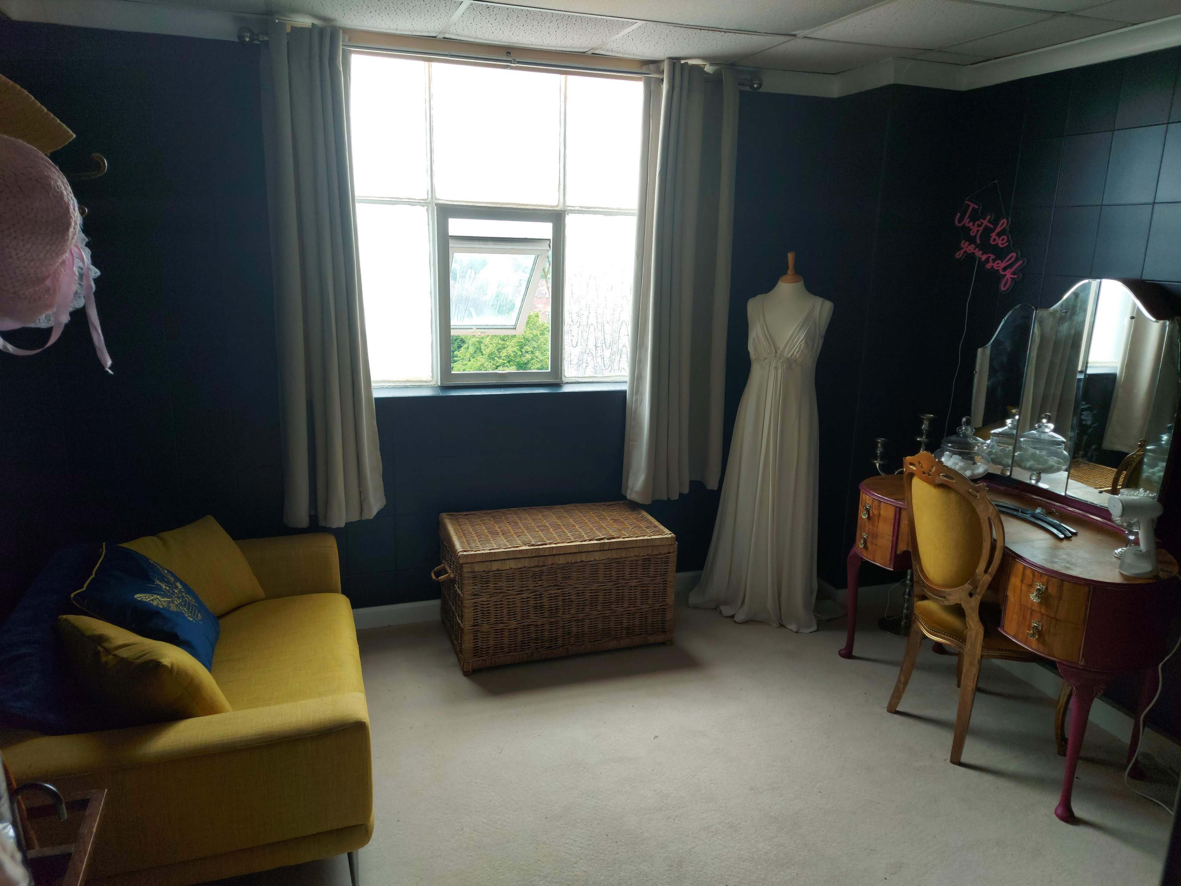 A small, dimly lit room features a yellow sofa, a wicker storage chest, a vintage dressing table with a mirror, and a mannequin displaying a white dress, all against dark walls and a window letting in natural light.