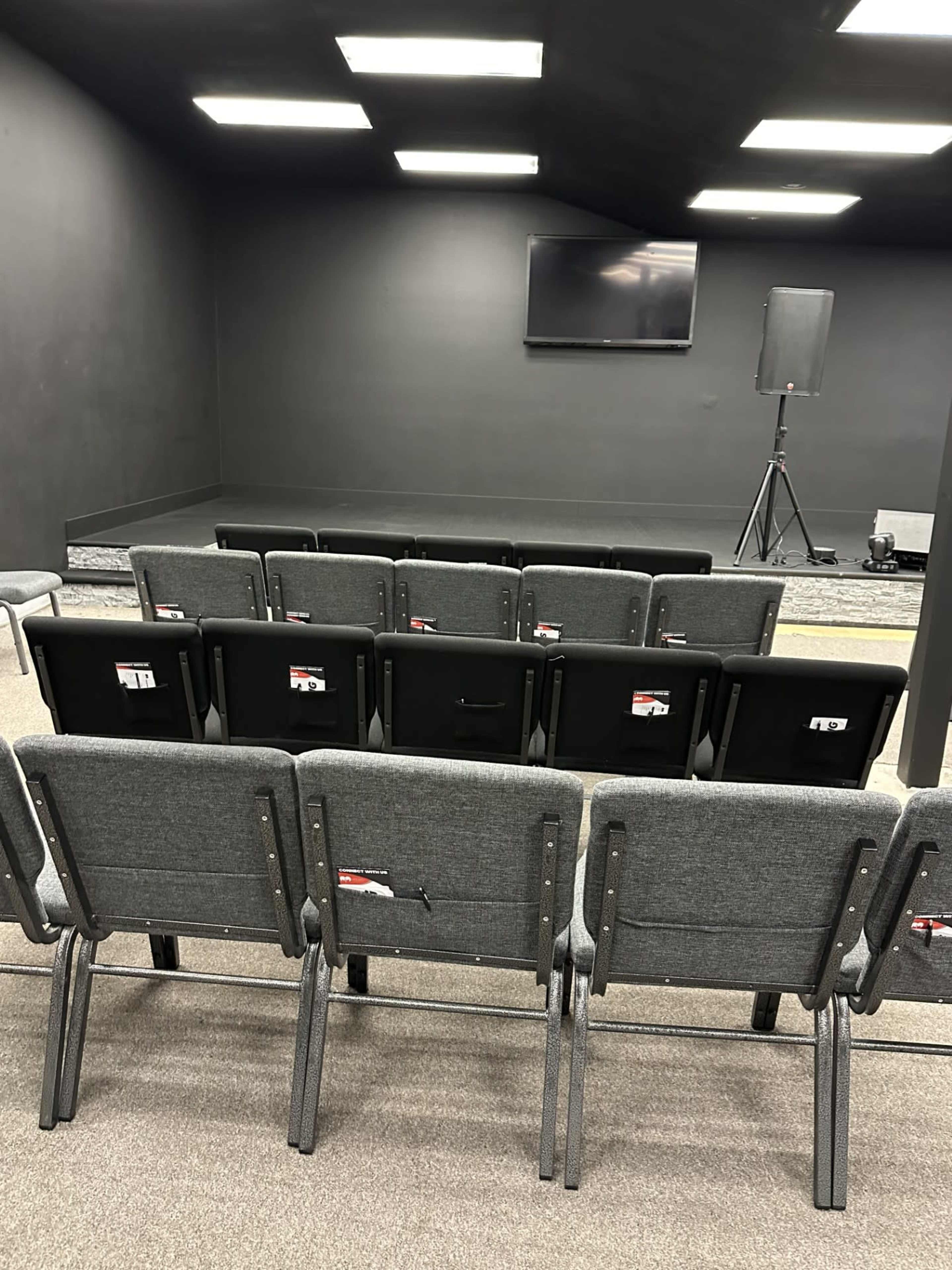 A row of gray chairs facing a small stage area with a screen and a speaker setup in a dimly lit room.