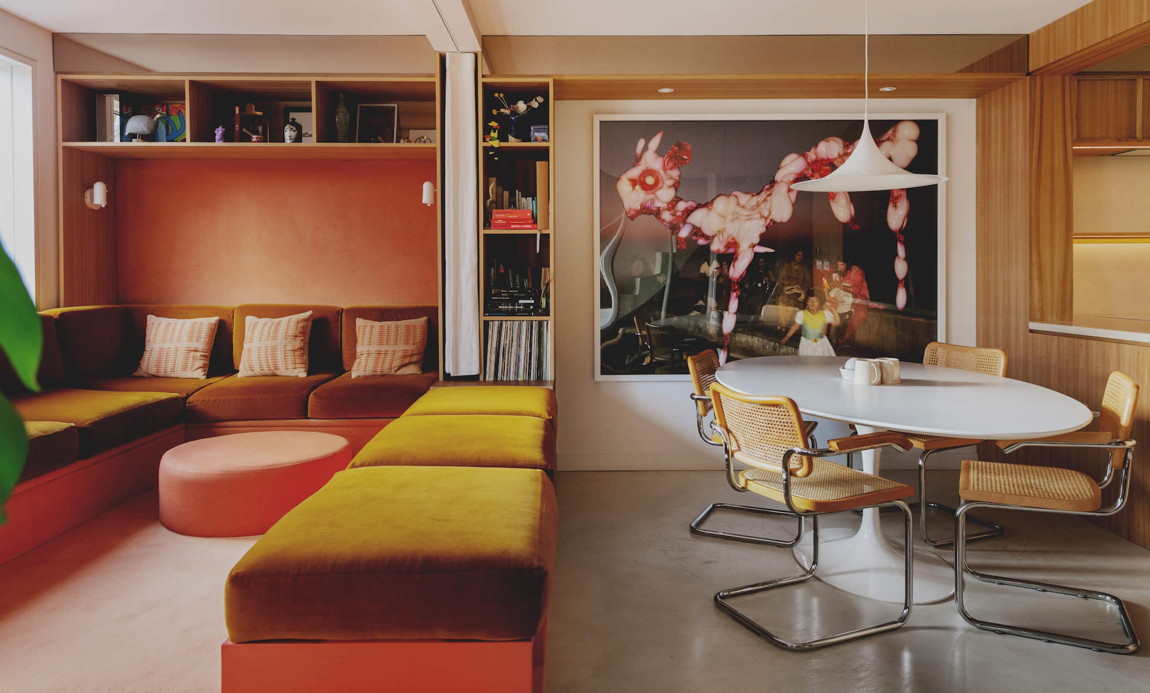 A cozy living space features a large, L-shaped sofa in earthy tones, a circular ottoman, and a round dining table surrounded by metal chairs, with a colorful artwork on the wall.