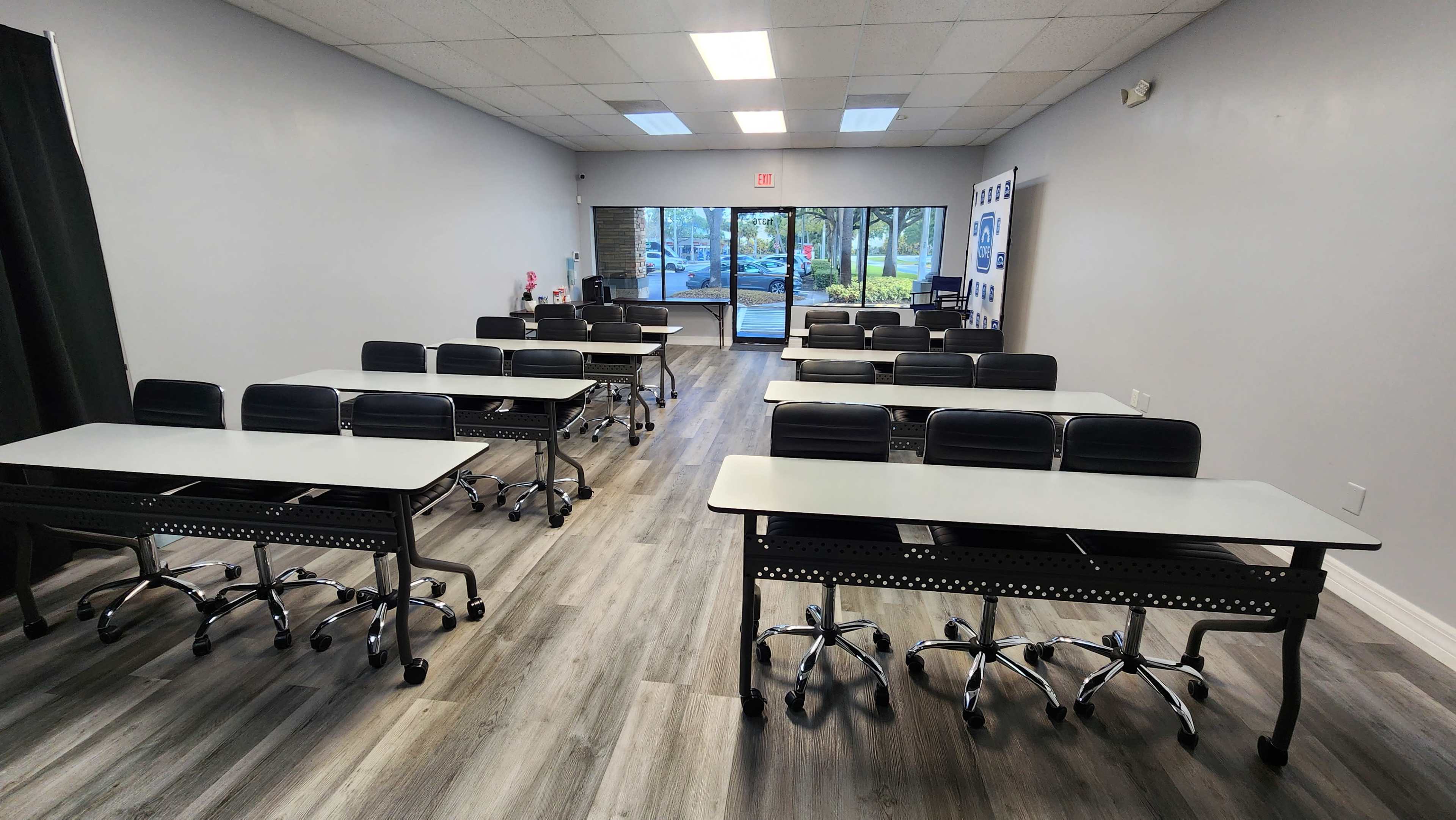 The image shows a brightly lit classroom with rows of steel-frame tables and black rolling chairs facing the entrance.