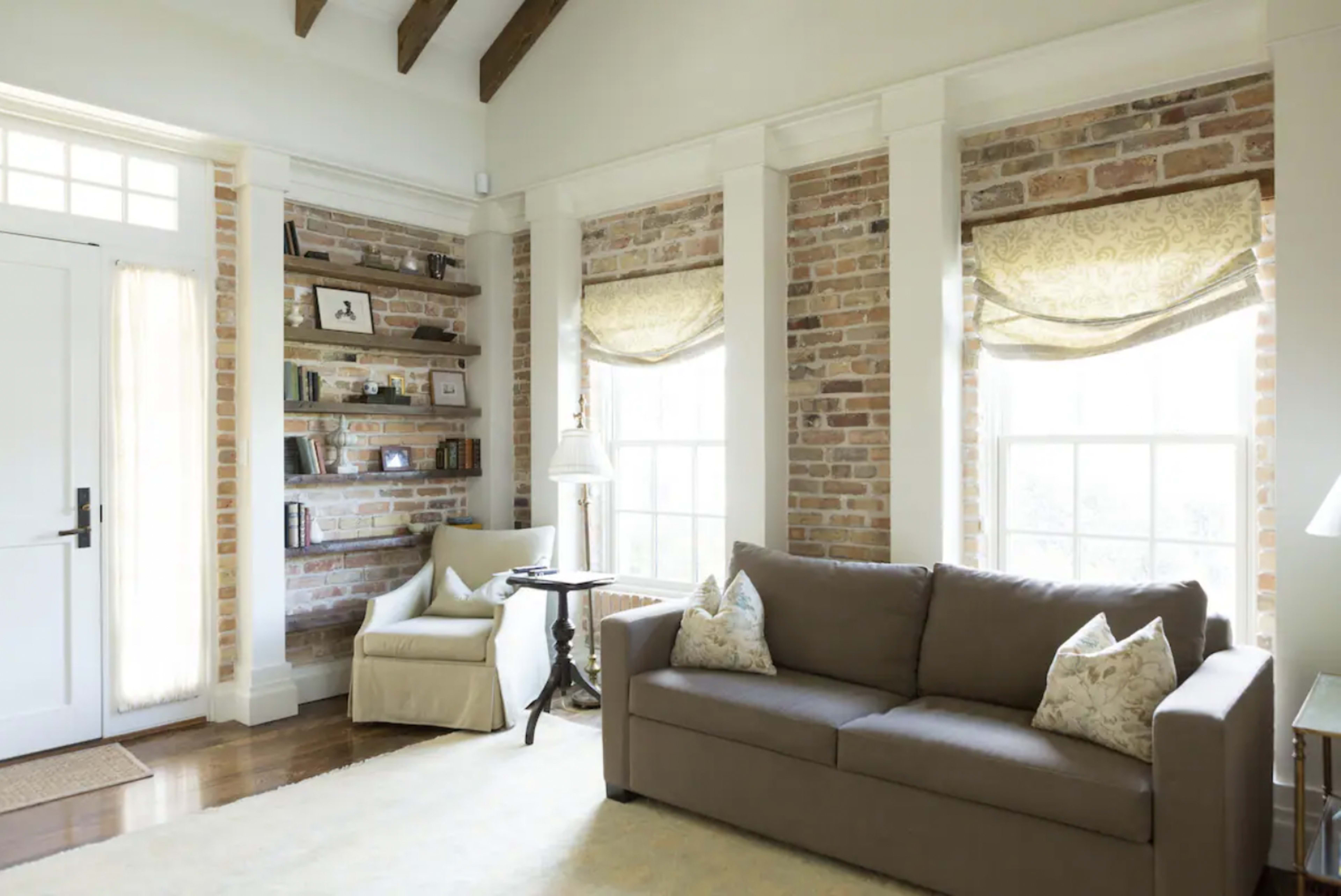 The living room features a brown sofa, a light-colored armchair, and exposed brick walls with large windows adorned with fabric shades.