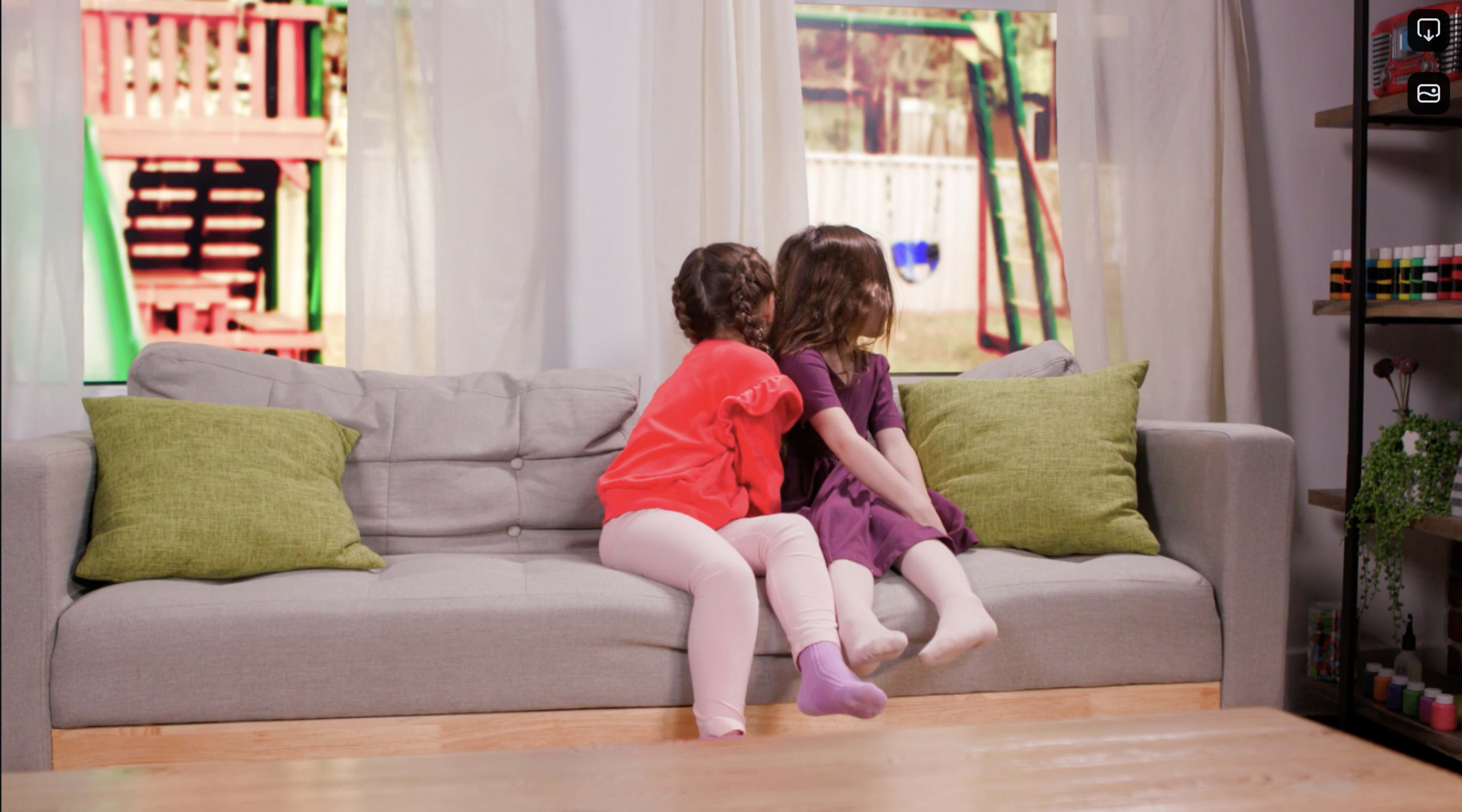 Two girls sit on a light gray couch, facing away from the camera, with one wearing a red top and the other in a purple dress, while colorful outdoor playground equipment is visible through the window behind them.