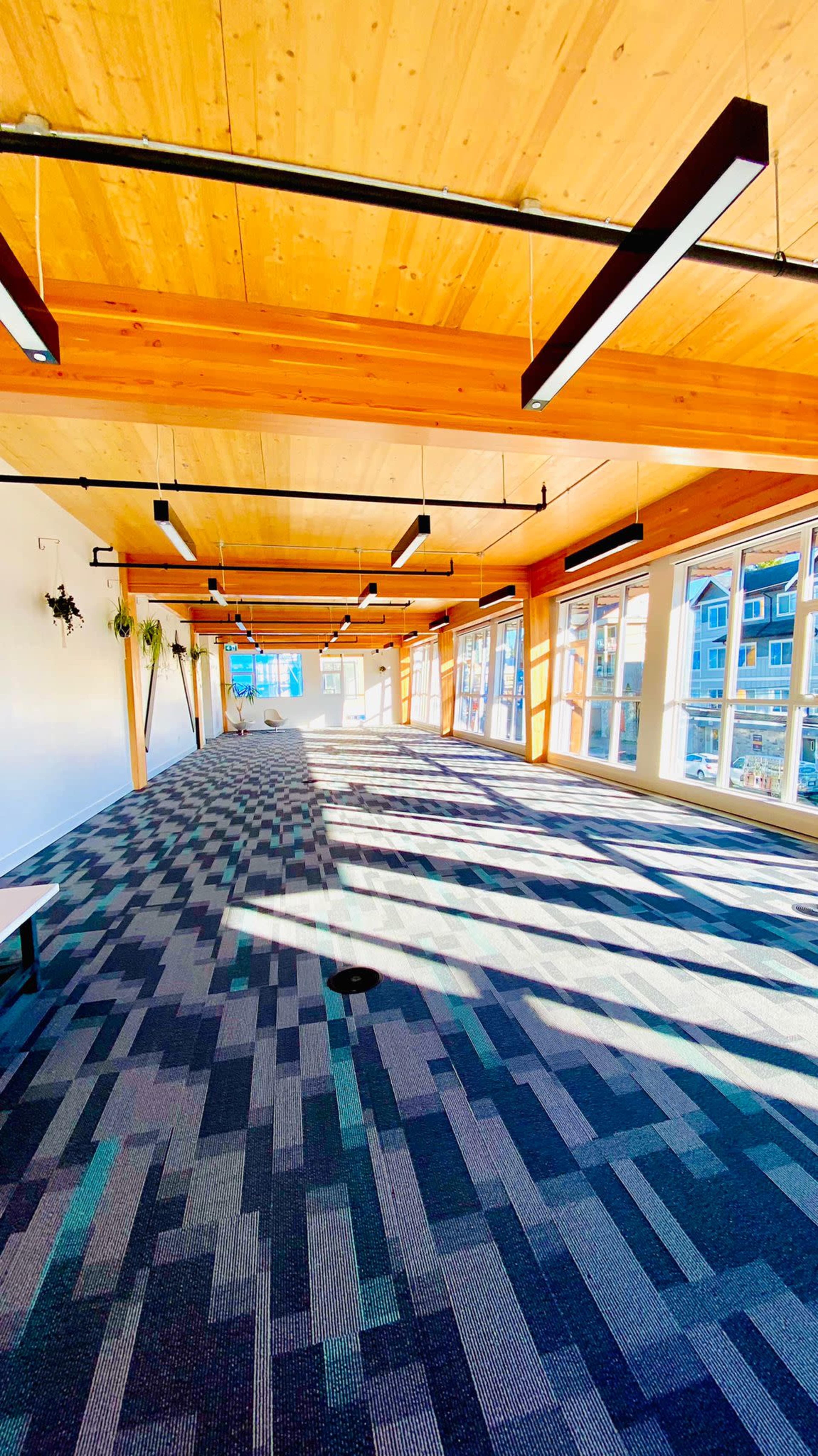 A spacious interior with wooden beams and a patterned carpet, illuminated by natural light streaming through large windows.